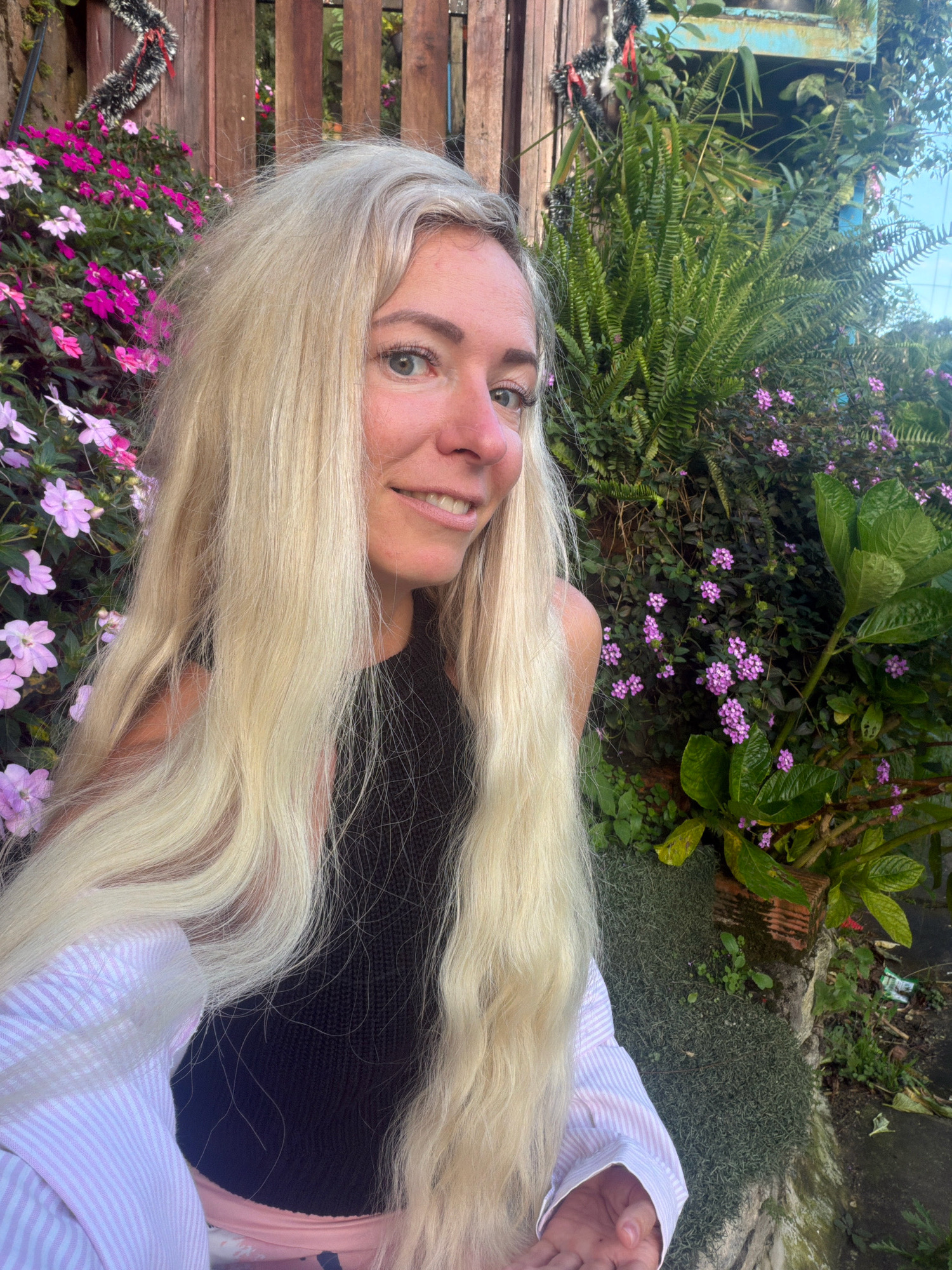 A young Caucasian woman with long, blonde hair smiles at the camera. She is wearing a black tank top and a pink and white striped button-up shirt. She is positioned in front of a lush garden with various plants and flowers, including pink impatiens, purple lantana, and ferns. In the background, a wooden fence is visible. The scene appears to be outdoors, possibly in a residential area or park in Xuân Thành, Vietnam. The lighting suggests it is daytime.