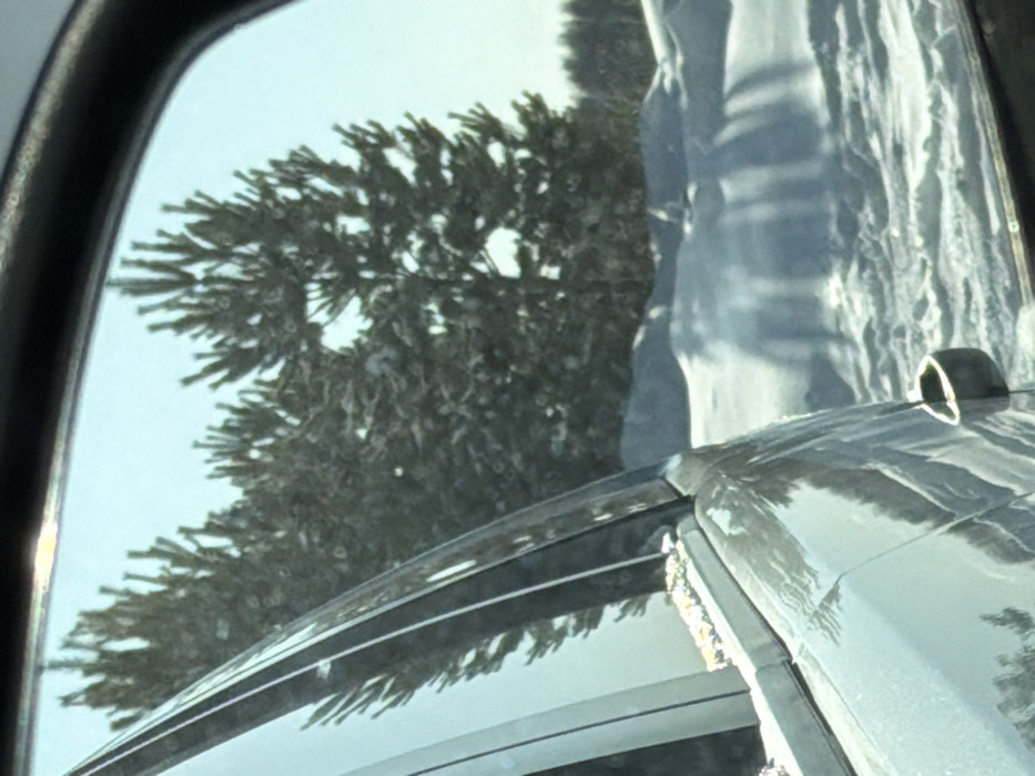 The image depicts a winter landscape viewed from the passenger side of a light-colored vehicle in Tyumen, Russia. On the left, a silver-grey vehicle's window frame, a portion of the A-pillar, and a small segment of the side mirror are visible. Small, yellow-orange granular debris is present on the vehicle's window seal. Through the vehicle's window and mirror, a snow-covered environment is observed. Several dense coniferous trees, heavily laden with snow, dominate the midground. The ground is entirely covered by deep, uneven snowdrifts, exhibiting long, distinct shadows cast by the trees, indicating direct sunlight. The sky overhead is clear and light blue. No individuals, animals, or other man-made structures are present in the external scene, depicting a natural, forested area.