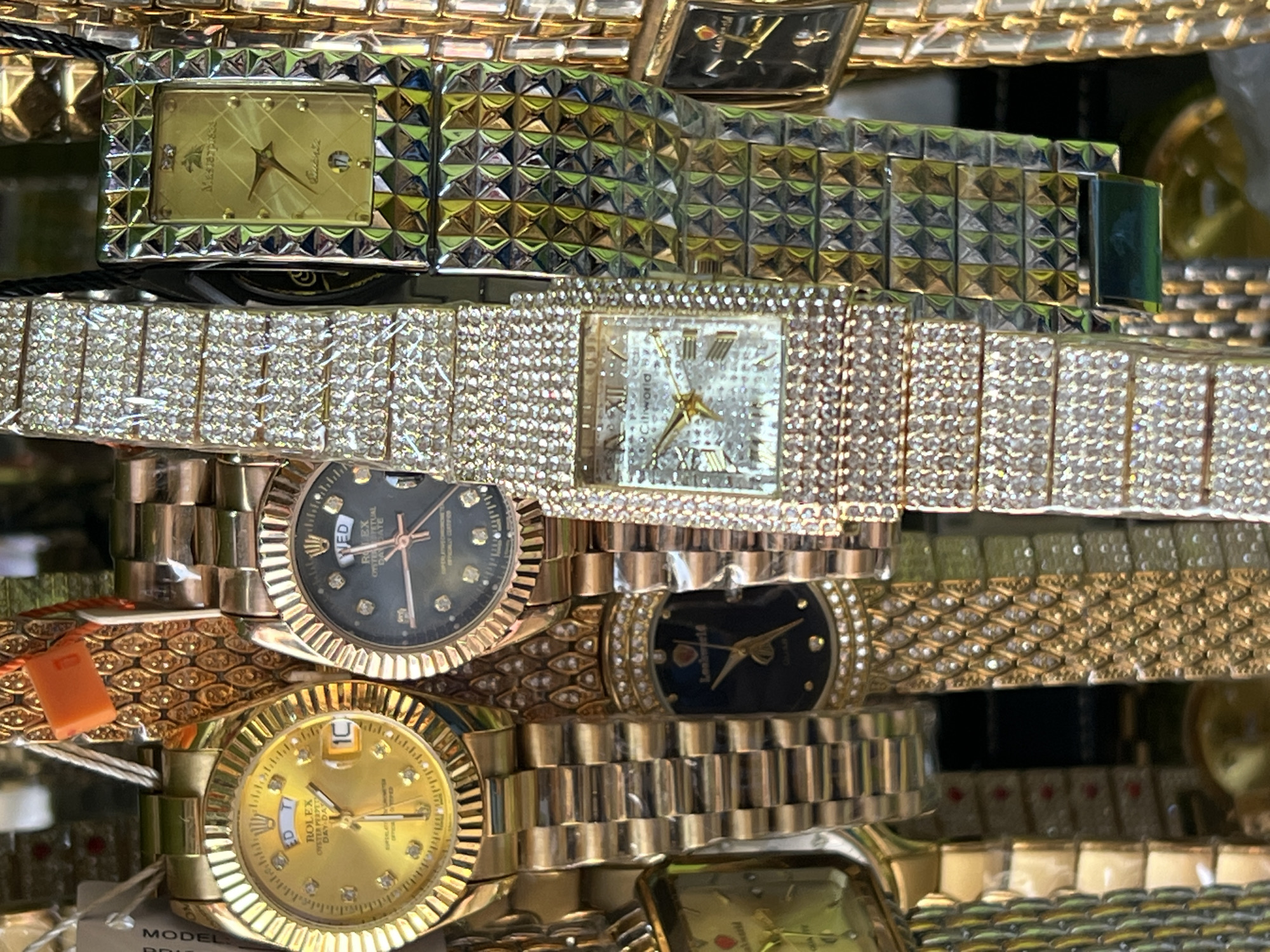 The image depicts a close-up, eye-level view of a display of numerous gold-toned watches. The watches are varied in design, with some featuring diamond-like embellishments on the face and band, while others have more minimalist dials. Several watches are layered over each other, creating a dense composition. The background is blurred and out of focus, suggesting a retail environment. The lighting is bright, reflecting off the metallic surfaces of the watches. The setting appears to be a market stall or a jewelry store. This scene is located in Bukuru, Nigeria.