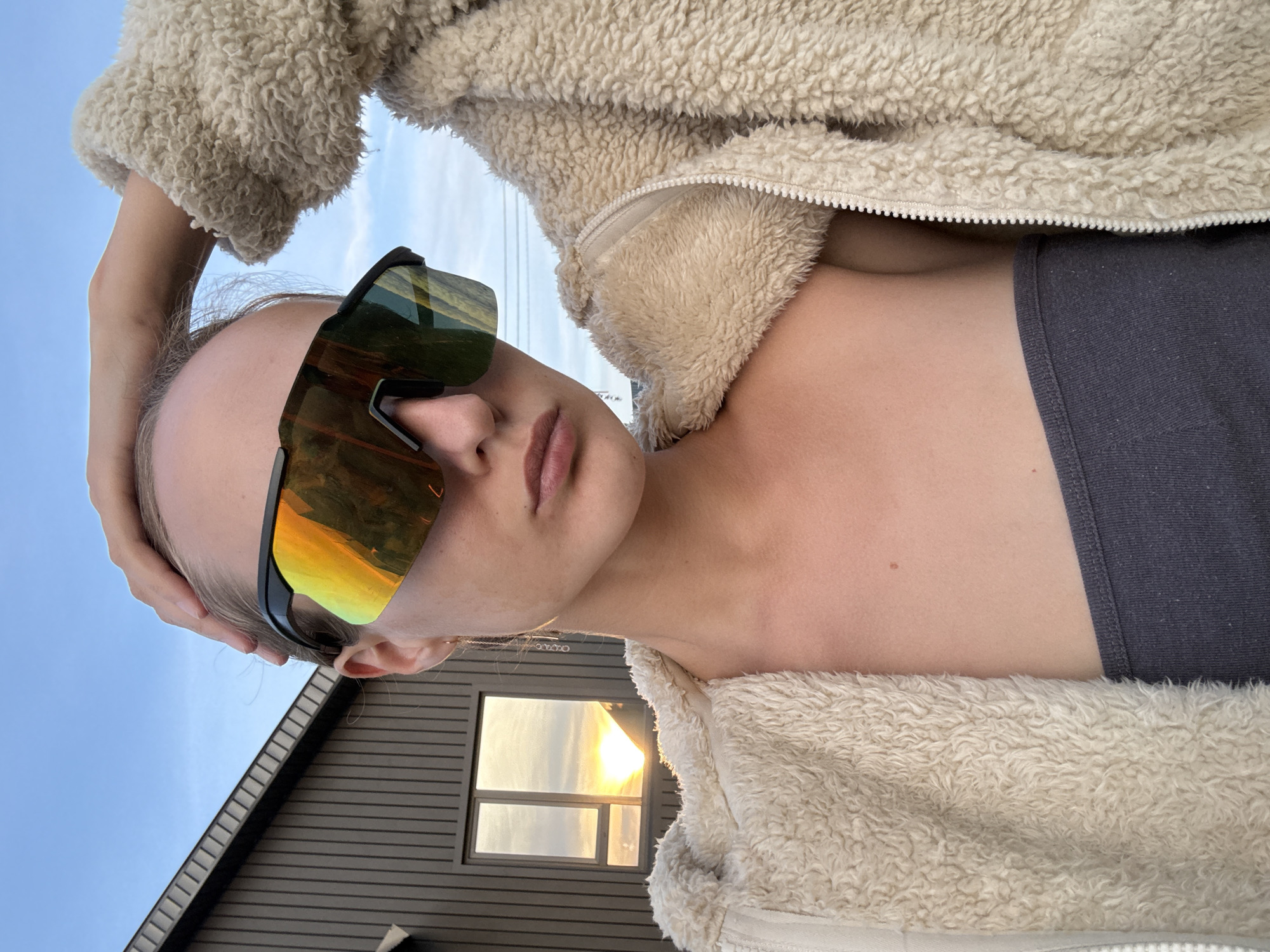A young woman, with her hand on her head, wears mirrored sunglasses and a fluffy beige jacket. She is standing in front of a modern, dark-brown house with a large window reflecting a sunset. The sky is a clear blue with a few wispy clouds. The scene appears to be in a rural or suburban setting, possibly in the village of Podshivalovo, Russia.