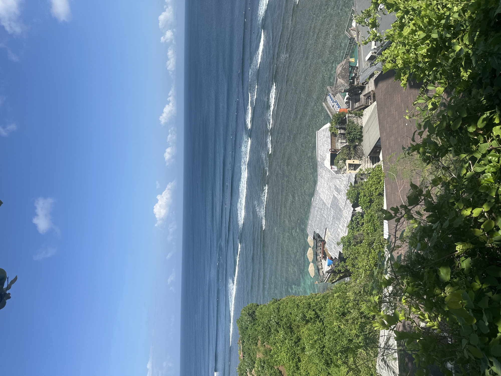 The image shows a coastal scene in Kuta Selatan, Indonesia, with a clear view of the ocean and sky. A line of waves breaks along the coastline, and several people are visible surfing in the water. On the shore, there is a structure with a roof, surrounded by lush green vegetation. The sky is blue with scattered clouds. The angle of the shot suggests it was taken from a high vantage point, likely a cliff or elevated structure.