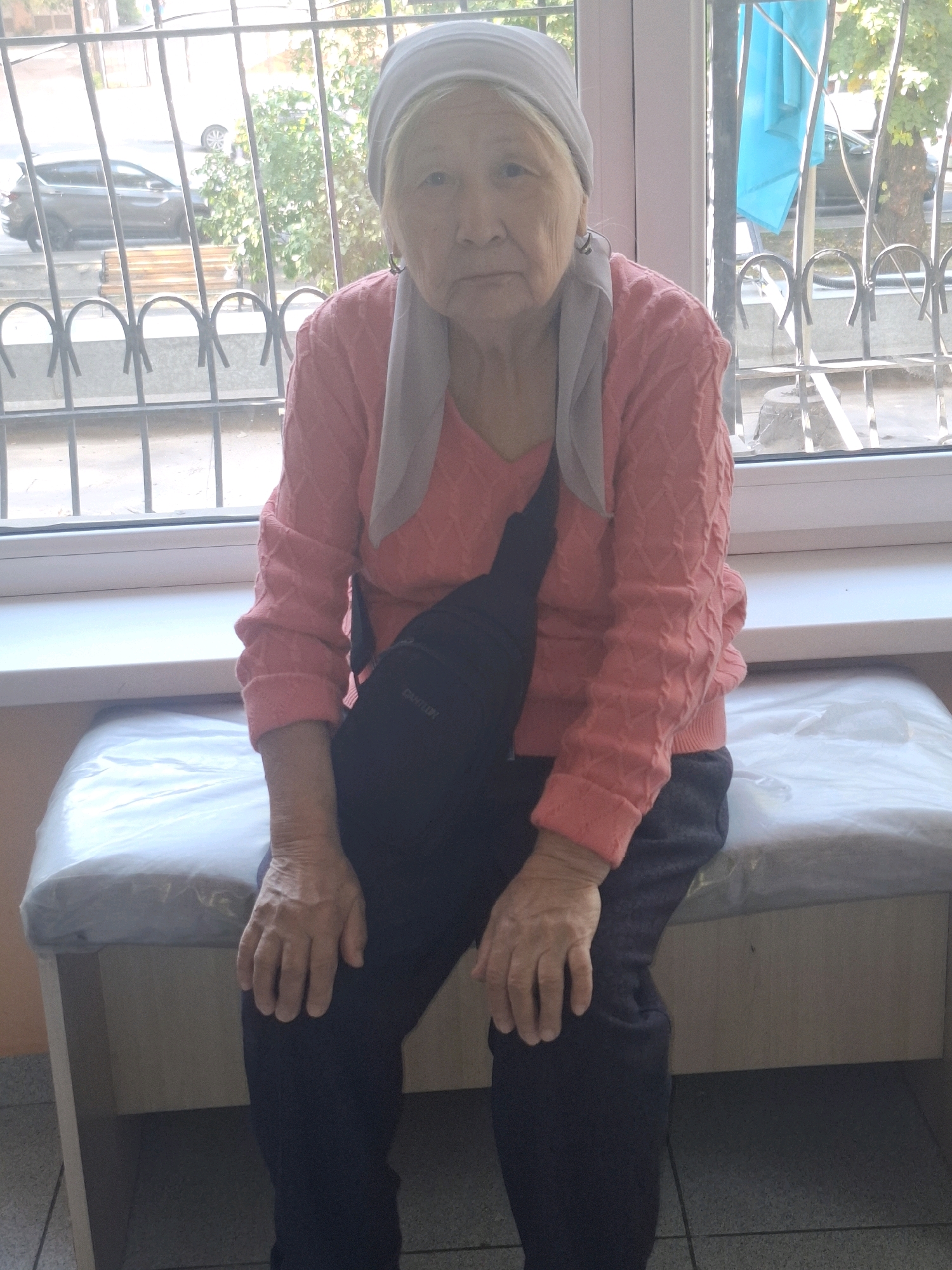 An elderly woman wearing a headscarf, a pink knitted sweater, and dark pants sits on a cushioned bench in front of a window with a metal grate. A dark crossbody bag is worn over her shoulder. Outside the window, a car and trees are visible. The room appears to be a waiting area, possibly in a medical facility, given the nature of the bench and the clinical-like plastic covering on it. The lighting suggests it is daytime. This scene is located in Almaty, Kazakhstan.