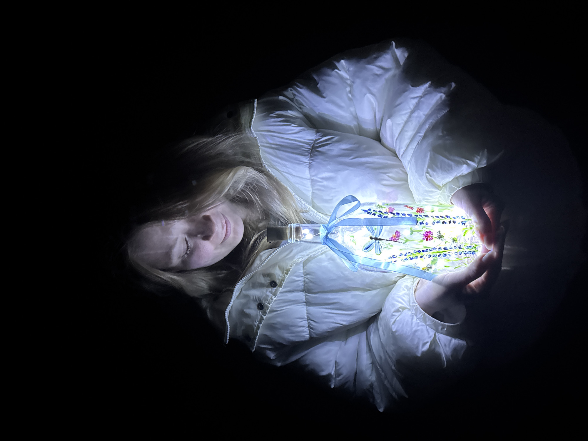 A woman with blond hair is holding a bottle with a blue ribbon and a dragonfly design on it. The bottle is illuminated from within, casting a bright glow on her hands and white puffy jacket. The background is dark, suggesting nighttime. There is no evidence to suggest the location is in Вассята, Russia.