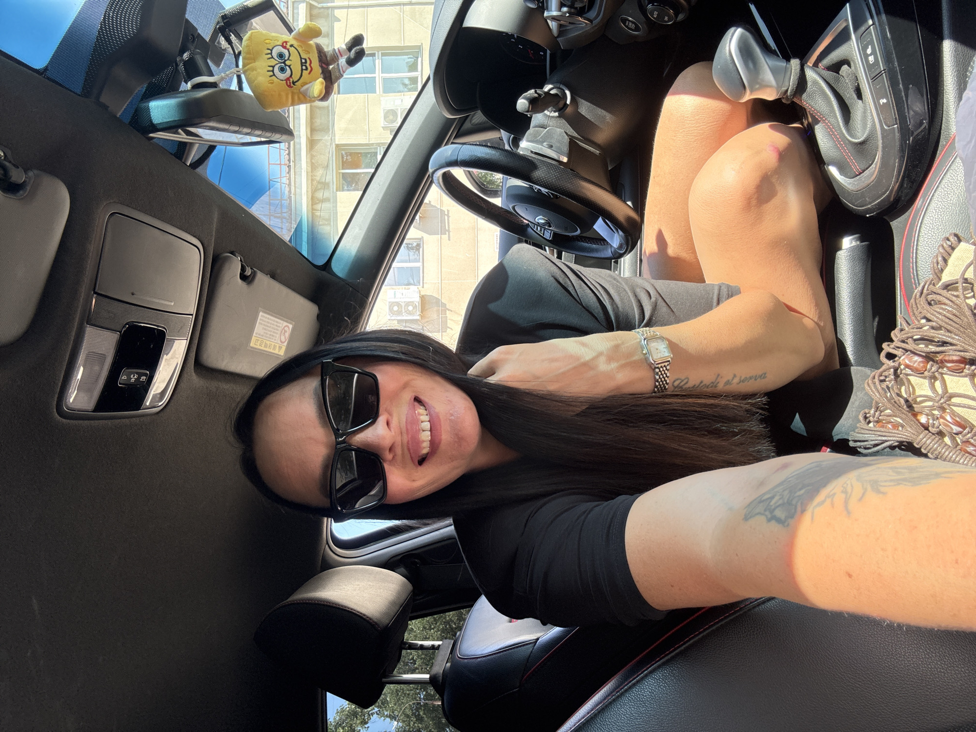 In this close-up selfie, a woman with long dark hair and sunglasses smiles as she sits in the driver's seat of a car. She is wearing a black top and is positioned so her right arm is resting on her leg, with a tattoo visible on her forearm. A watch is on her left wrist. The car's interior is visible, including the steering wheel, gear shift, and a Spongebob Squarepants plush toy hanging from the rearview mirror. Outside the car, a building with windows and air conditioning units can be seen, bathed in sunlight. The environment suggests an urban setting, likely Chaikovsky, Russia, given the presence of typical Russian apartment buildings and the style of air conditioning units. The image captures a casual moment, possibly a selfie taken while parked.