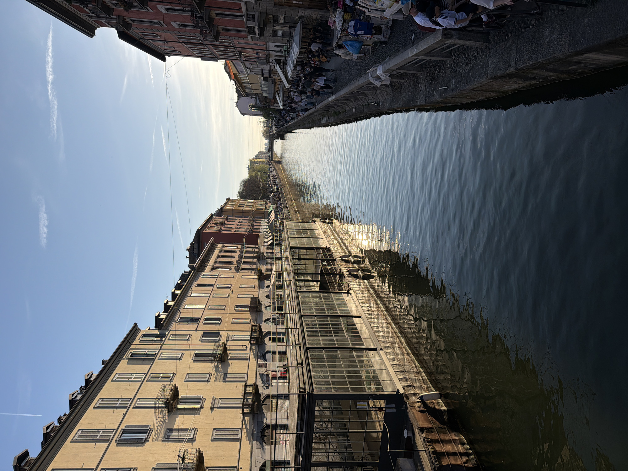The image captures a canal scene in Milan, Italy. The waterway stretches into the distance, flanked by buildings on either side. The water reflects the surrounding structures and sunlight. On one bank, tables and chairs are set up, occupied by people who appear to be dining or socializing. The buildings vary in color and architectural style, ranging from beige to red, with multiple windows and some balconies. The sky is visible above the buildings, showcasing a clear blue expanse with some contrails. Overall, the scene depicts a vibrant urban environment along the Navigli canals of Milan.