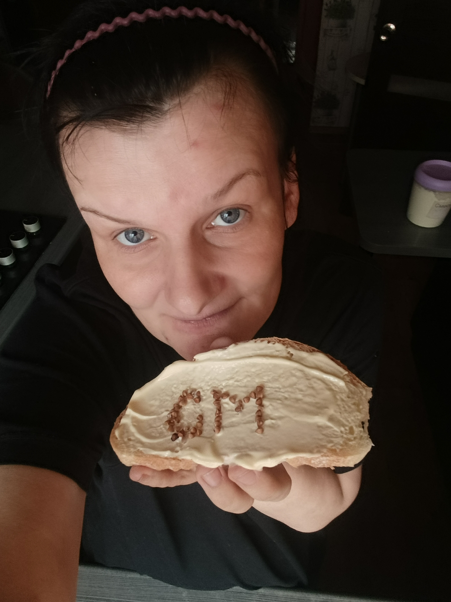 A high-angle selfie shows a fair-skinned person with blue eyes, dark hair pulled back with a pink headband, holding a slice of bread with a creamy spread. The spread is decorated with rows of small, brown items resembling seeds or grains, spelling out the letters "OM". The person wears a black t-shirt. In the blurred background, a stove with black knobs, a small container with a lavender lid, and a decorative cabinet with plants on the doors are visible. The location in the city of Фоки, Russia is not deducible from the image alone.