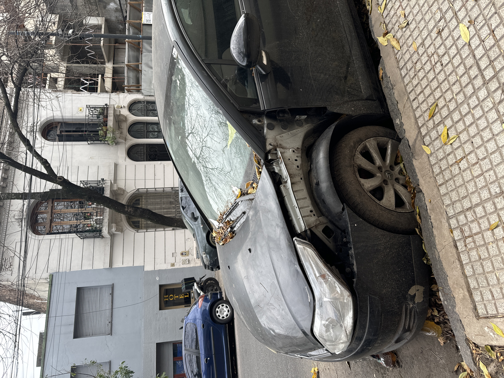 The image depicts the front of a black car parked on a sidewalk in Buenos Aires, Argentina. The car has significant damage to its front end, with a missing bumper and exposed internal structure. The front tire is pressed against the curb. The street is lined with buildings, including one with arched windows and another that appears to be a hotel. There are fallen leaves scattered around the car and on the sidewalk, indicating that it's autumn or winter. Other vehicles, including a blue car, can be seen parked behind the damaged car.