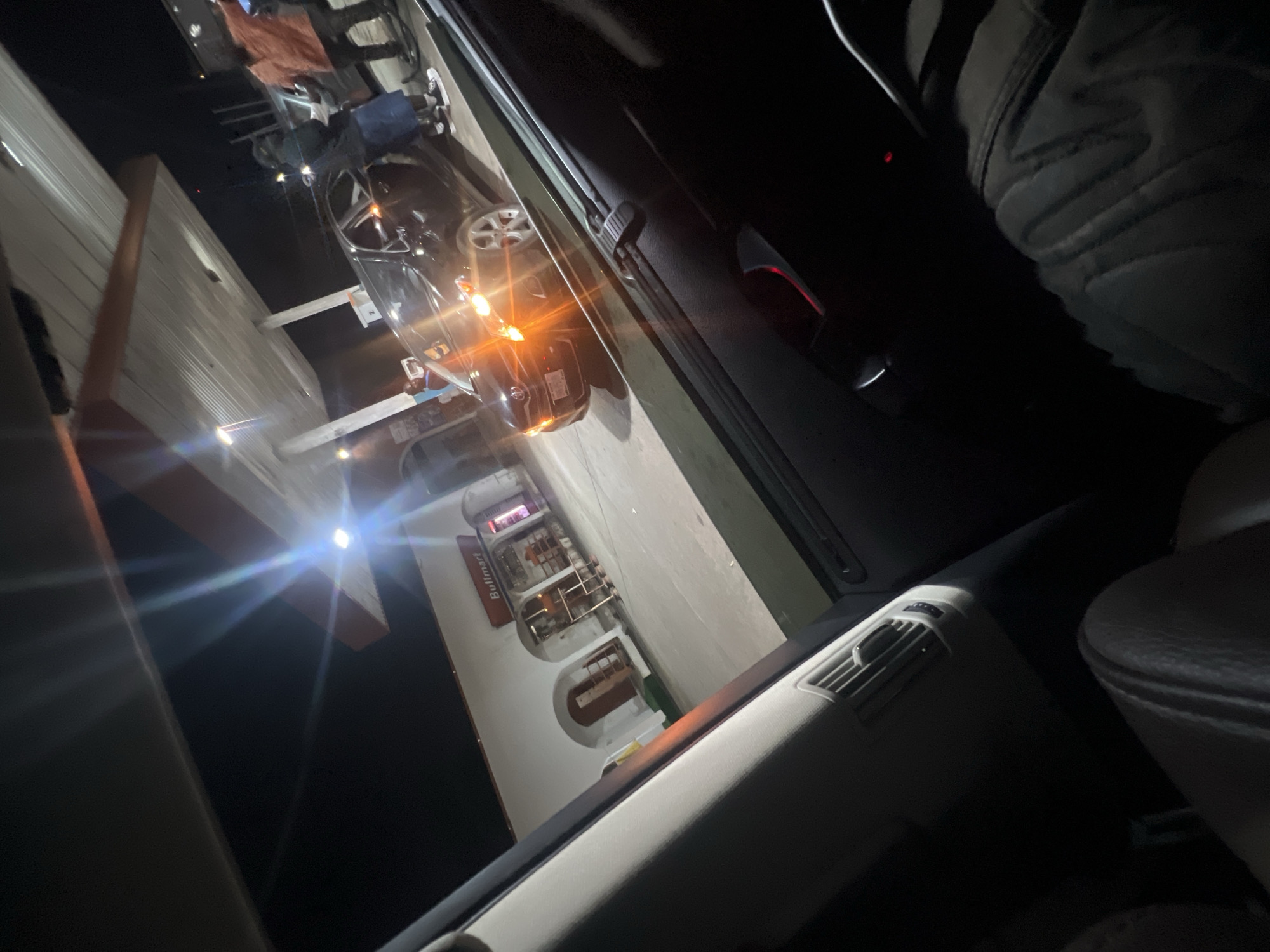 The image shows the interior of a car parked at a Bulkmart store at night. The Bulkmart signage is visible on the storefront. Another car is parked in front, its headlights illuminating the scene. The interior of the car includes a partial view of the driver's seat, the passenger side air vent, and the car door. The outside environment appears dark and urban. Due to the fact Bulkmart store is in Abuja, Nigeria it can be said the location is Abuja, Nigeria.