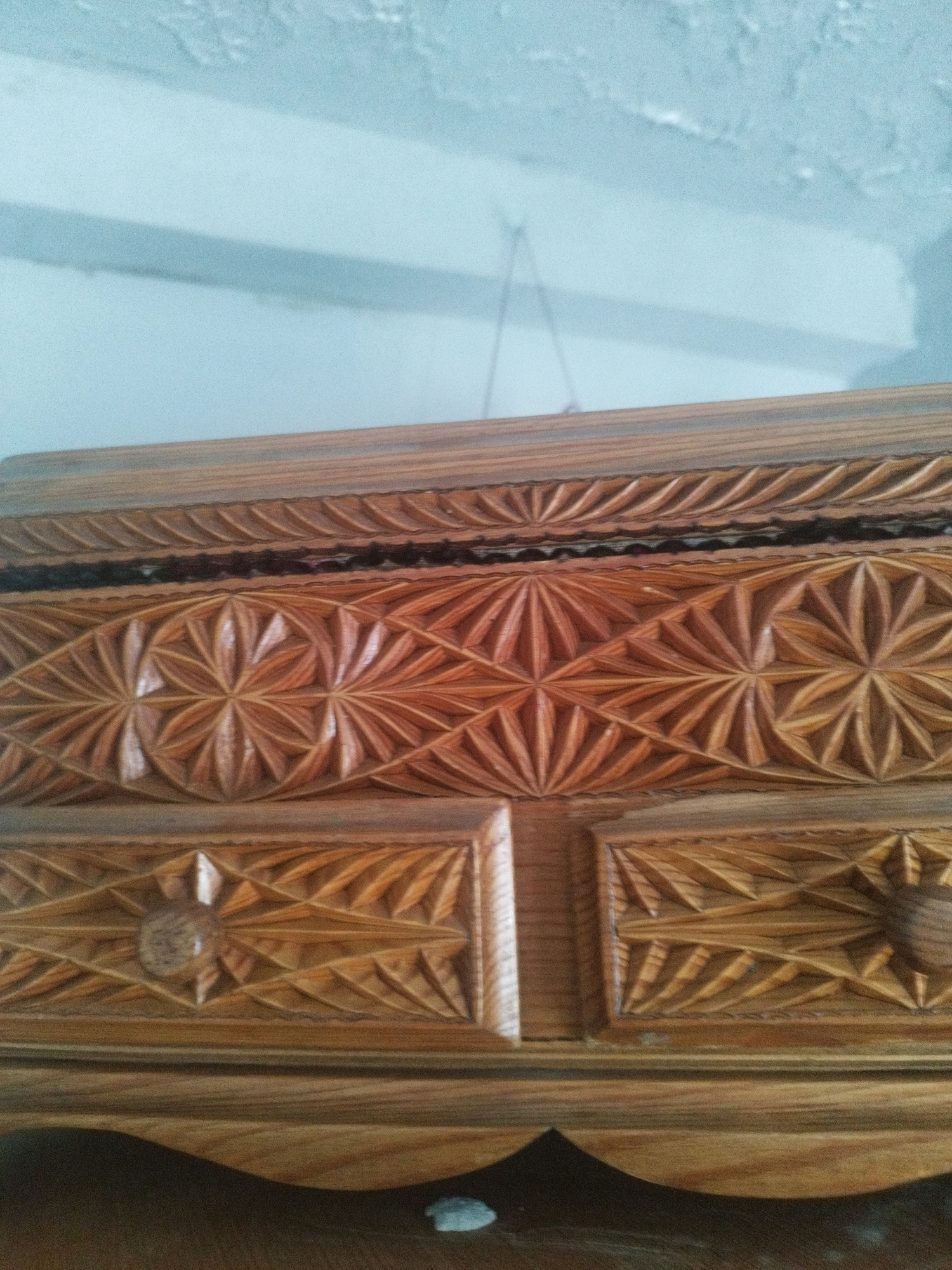 A close-up, eye-level, interior shot shows the carved wooden surface of what appears to be a piece of furniture, possibly a cabinet or chest. The wood is a warm, medium brown with visible grain. The surface features intricate geometric carvings arranged in bands. The top band has a repeating pattern of stylized leaf or fan shapes. Below this is a wider band with a repeating starburst pattern created by radial and triangular cuts. The bottom visible band has a diamond pattern with triangular elements. There are also two rectangular drawer fronts visible, each with a central starburst design and a surrounding diamond pattern. The background is out of focus, showing a light blue wall and a thin wire hanging from the ceiling. The lighting appears to be natural, coming from the right. The scene is static and offers no information about the city of Oskemen, Kazakhstan.