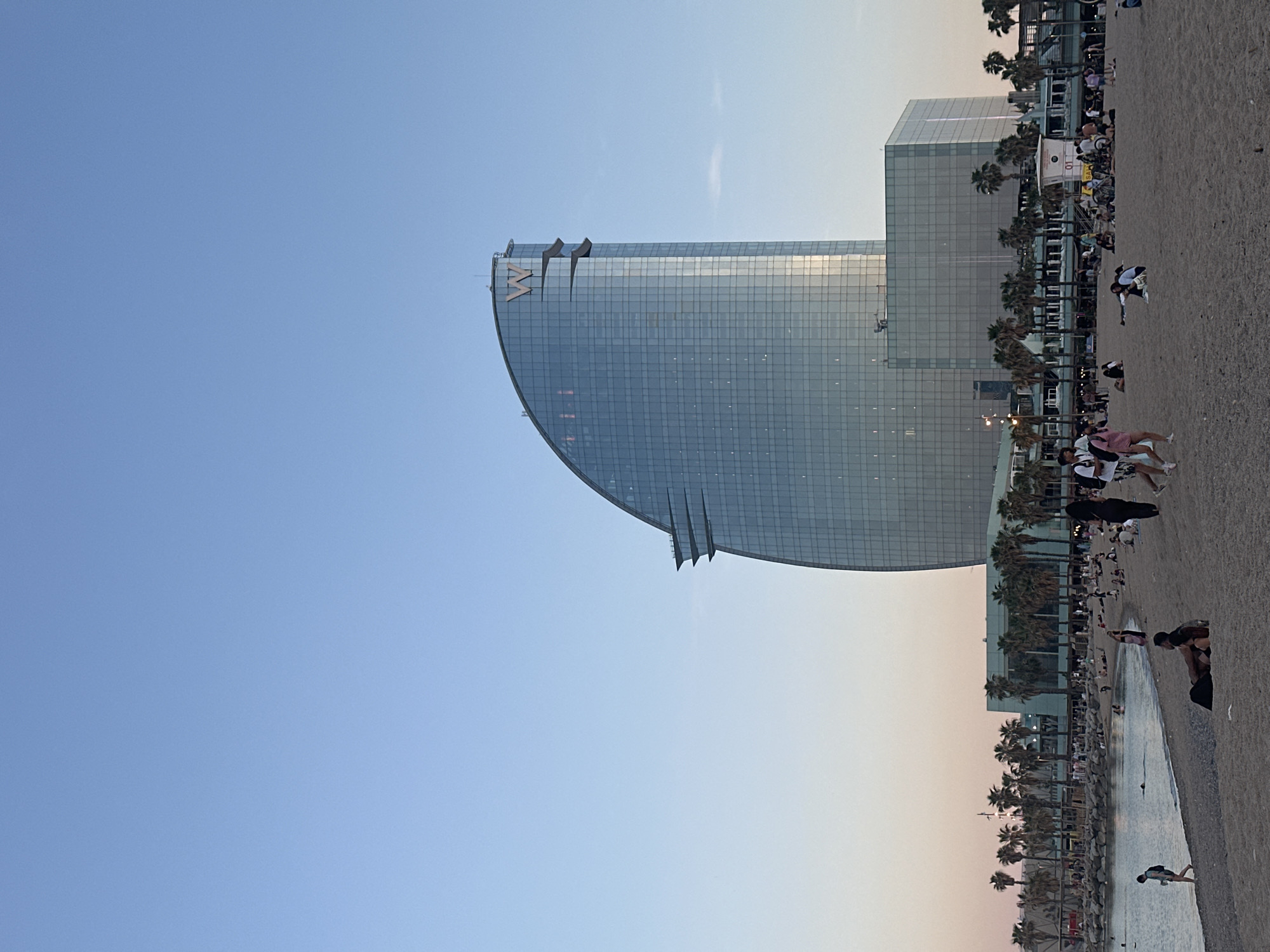 The image depicts the W Barcelona hotel, a distinctive sail-shaped structure, situated along the coastline in Barcelona, Spain. The hotel is a modern, glass-clad building with the "W" logo prominently displayed on its curved facade. Adjacent to the hotel is a beach with palm trees lining the edge. There are people scattered along the beach, some appearing to be walking along the shoreline, others standing or sitting. The sky transitions from a light blue to a subtle gradient of orange and pink near the horizon.
