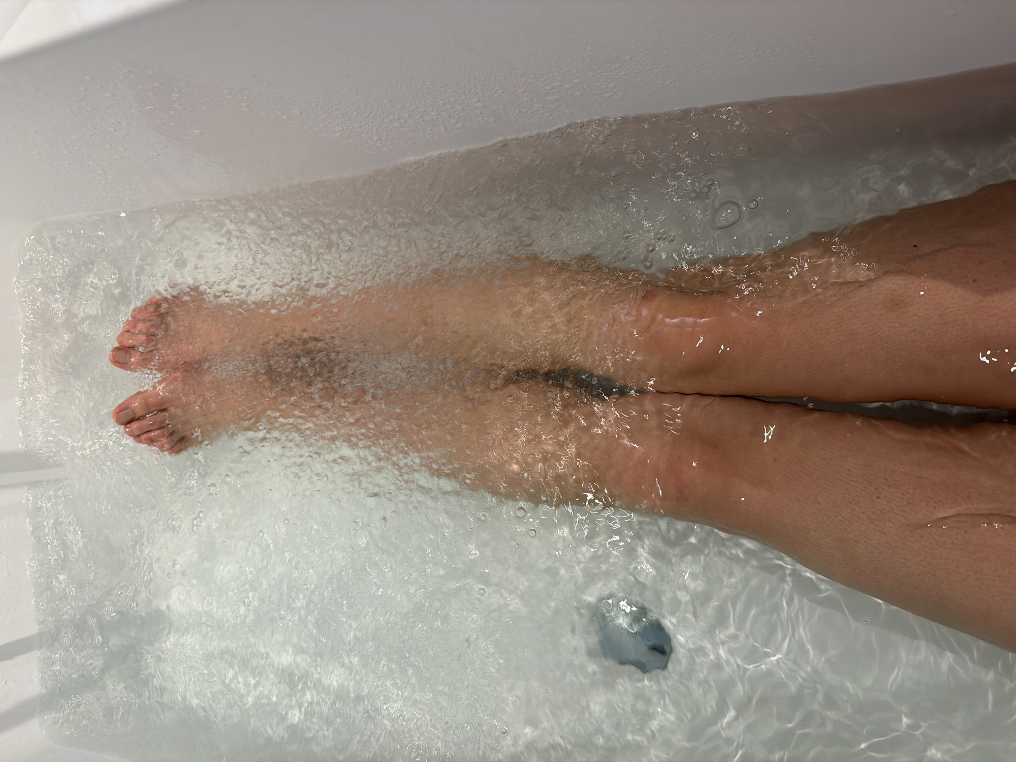 A top-down shot shows two legs submerged in a bathtub filled with clear water. The legs appear to be of an adult, with the feet visible on the left side of the frame. The water is slightly turbulent, creating ripples and bubbles that obscure the legs. The bathtub is white and appears to be made of ceramic or a similar material. A drain is visible at the bottom of the tub. The location of Mandelieu-la-Napoule, France cannot be determined from the image.
