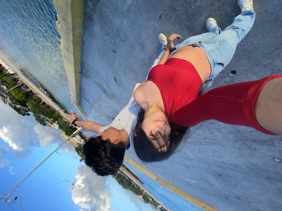 A selfie captures a young couple lying on a concrete surface beside water. The woman, wearing a red top and jeans, looks at the camera. A male figure lies beside her, wearing a light shirt. Buildings and trees line the waterfront in the background, and the sky is partially visible with scattered clouds.