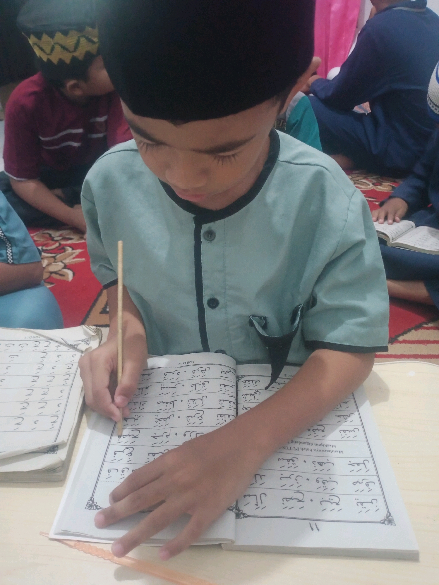 In a dimly lit room, a young boy wearing a light blue garment and a black skullcap focuses intently on an open book. He holds a thin wooden stick, likely a qalam, to point at the Arabic script within the book. Another open book lies to his left, with pages containing rows of Arabic characters. In the background, two other individuals, also wearing skullcaps, are visible. One is wearing a maroon shirt with white stripes, and the other is in a dark blue garment. They are seated on a red rug with an ornate pattern. The scene suggests an educational setting, possibly a Quranic class or religious study session. The room appears to be a domestic space, possibly a house in Bandar Lampung, Indonesia, given the cultural context implied by the attire and the activity.