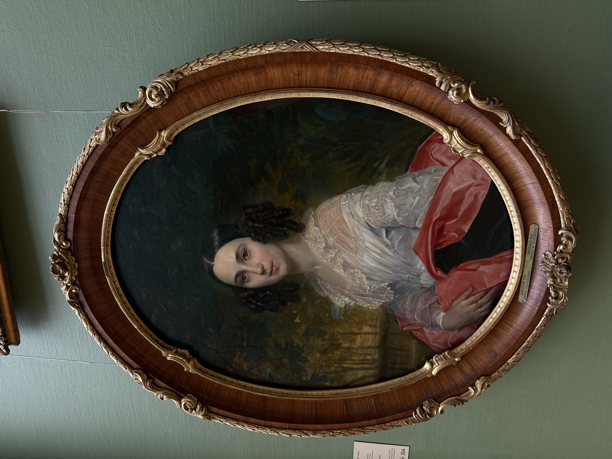 A framed oval portrait of a woman hangs on a green wall. The woman has dark hair, fair skin, and wears a white dress with lace details and a red shawl. She is looking directly at the viewer. The background of the portrait is a dark, indistinct landscape. The ornate frame is made of wood with gold accents. A small label is affixed to the right side of the frame. Another label is on the wall below the painting. Based on the writing on the label on the frame, it is very likely that this painting is currently located in Moscow, Russia.