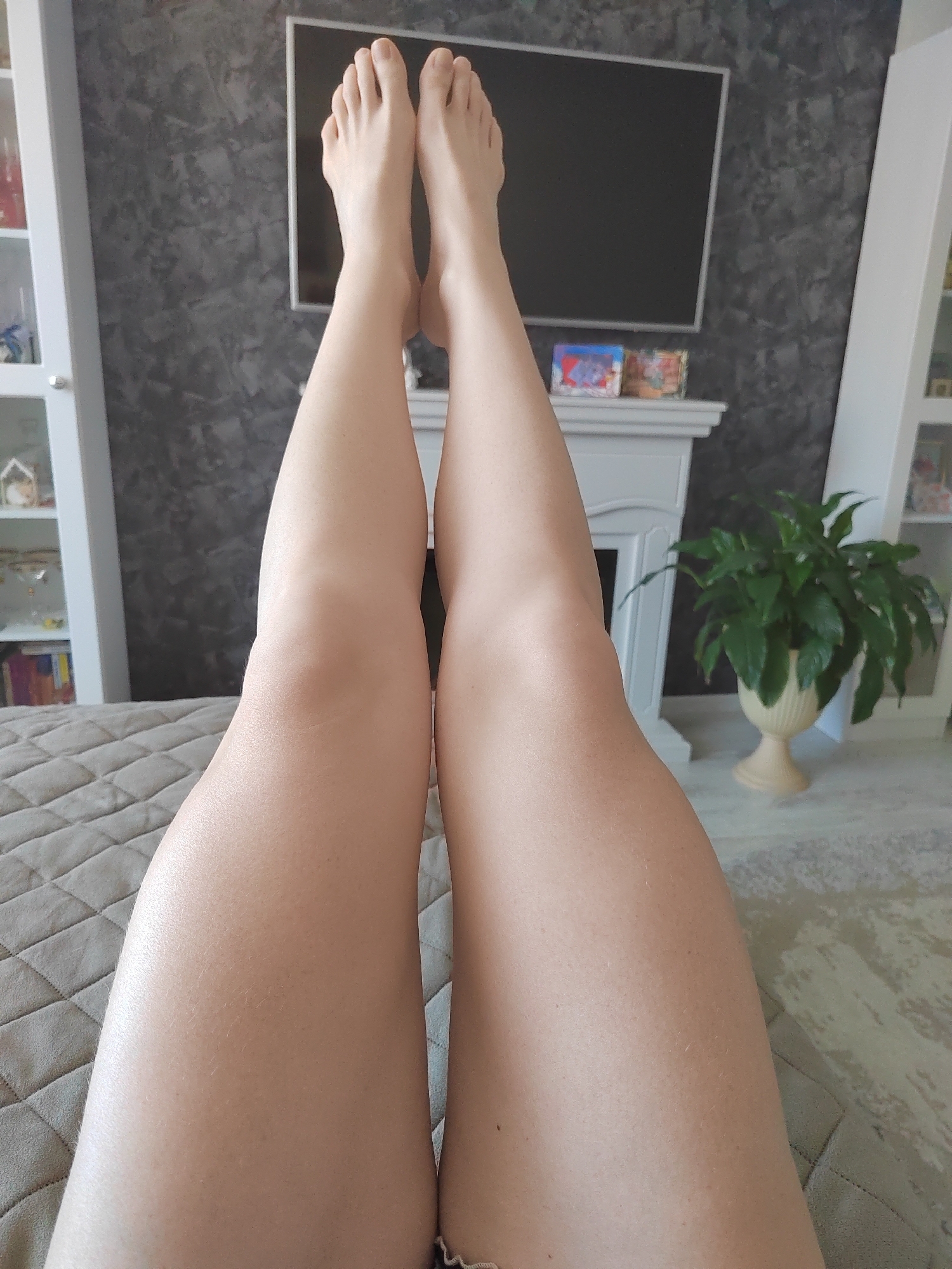 A first-person perspective shot shows a pair of bare legs and feet resting on a quilted bedspread. The legs are smooth and tanned, with some fine blonde hairs visible. The feet are positioned upward, with toes neatly aligned. In the background, a dark, textured wall serves as a backdrop for a television screen. Below the TV, a white fireplace mantle is adorned with small decorative items, including what appear to be miniature books or boxes. To the right of the mantle, a lush green plant in a cream-colored pot adds a touch of nature to the indoor setting. On either side of the mantle, white shelving units with glass doors are partially visible, suggesting a room with decorative storage. The overall lighting suggests daytime. The setting appears to be a living room or bedroom in a home, potentially in Saint Petersburg, Russia, given the location context. There is no discernible text in the image.