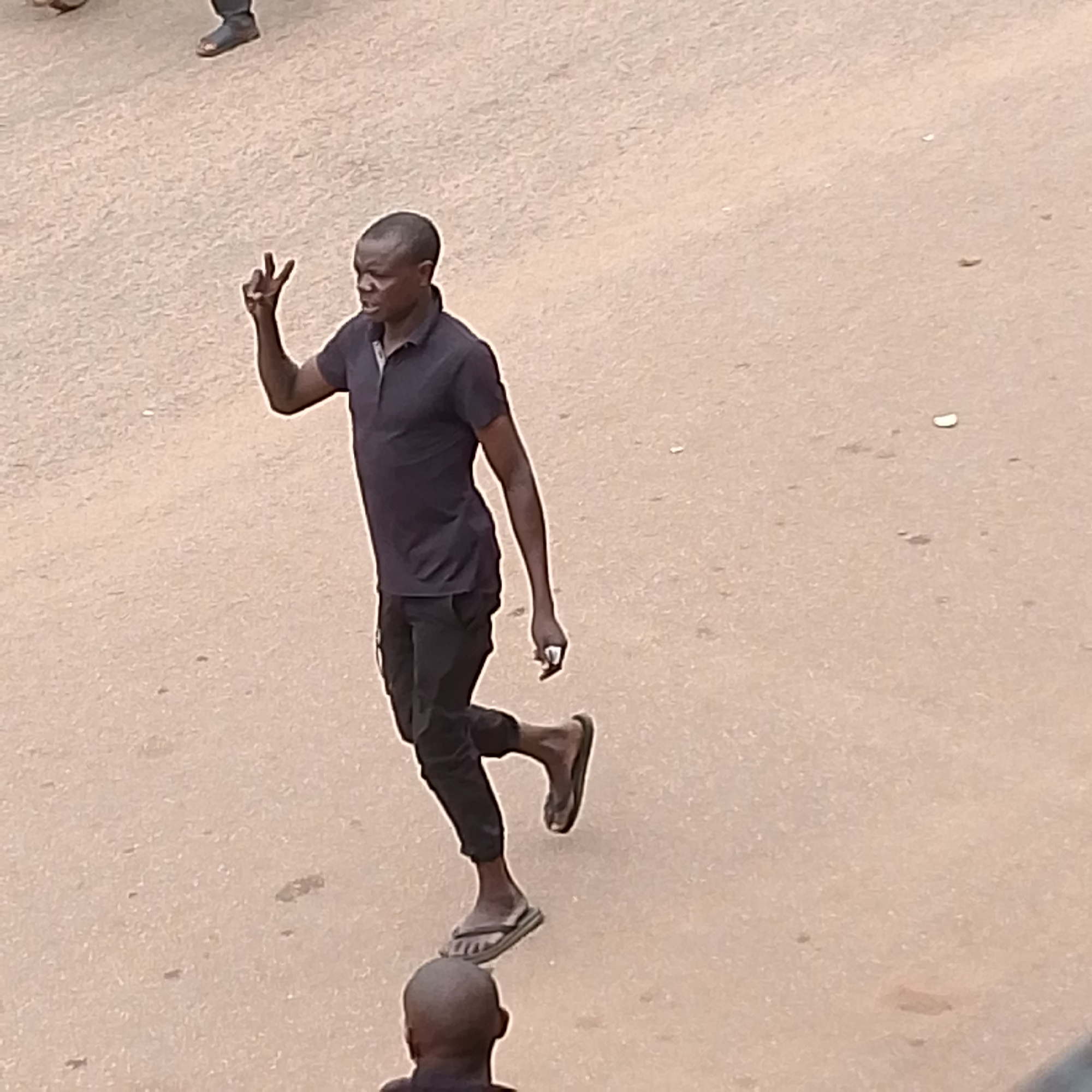 In an outdoor setting, a man in a dark blue polo shirt and dark pants is captured from a high angle, walking on a light brown, dusty surface. He is making a peace sign with his right hand. His left hand is down by his side. He is wearing dark flip-flops. Another person, whose head and shoulders are visible from the back, is in the foreground below the main subject. The ground is mostly flat and appears to be a road or open area. The image does not contain any identifiable landmarks or details that would suggest a specific location within Chaikovsky, Russia.