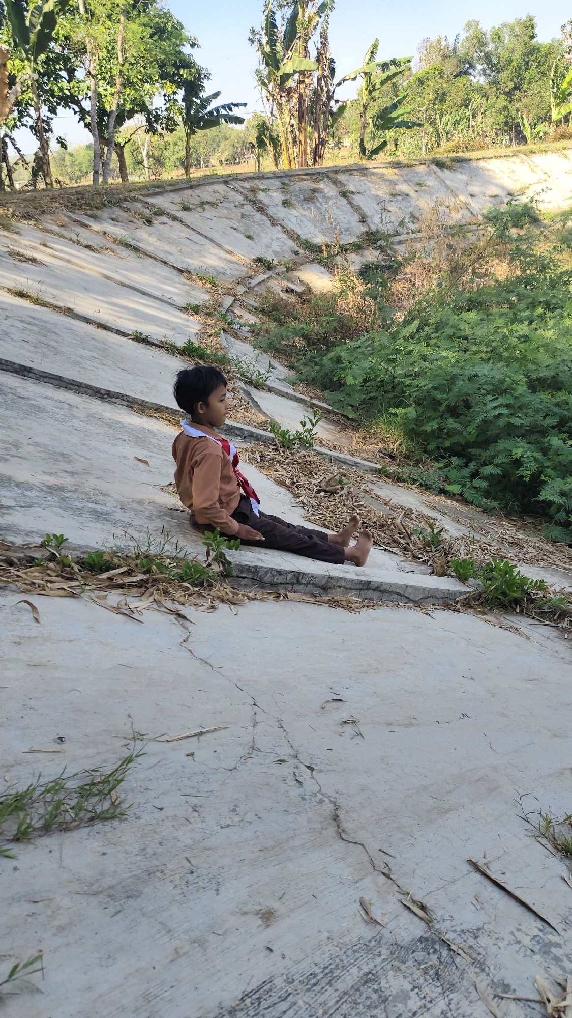 A young boy wearing a brown shirt and dark pants sits on a concrete embankment in Banyubunih, Indonesia. The boy is barefoot and appears to be looking to his right. The embankment is lined with concrete slabs and has sparse vegetation growing on it. In the background, there are trees and foliage. The sky is clear and blue. The overall scene suggests a sunny day in a rural or semi-rural area.