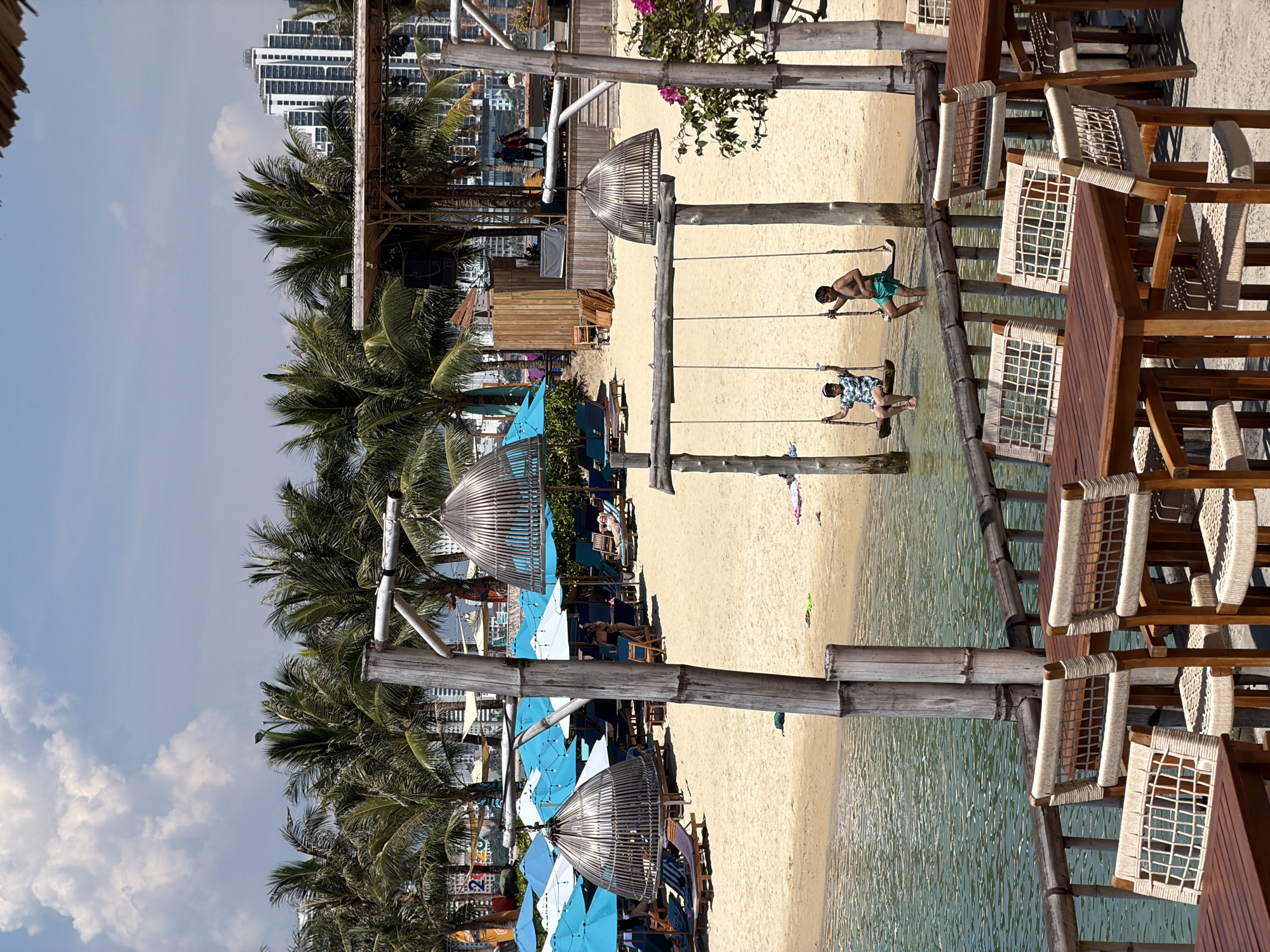 The image captures a coastal scene, possibly in Bac Nha Trang Ward, Vietnam, indicated by the presence of modern high-rise buildings in the background along with tropical features like palm trees. The foreground reveals a sandy beach gently sloping into the water, with two individuals enjoying rope swings suspended from wooden structures. Beach umbrellas and loungers suggest a recreational or resort area. Adjacent to the swings are wooden tables and chairs, seemingly part of a beachfront establishment. The environment is characteristic of a tropical coastal setting, reflecting a tourist destination.