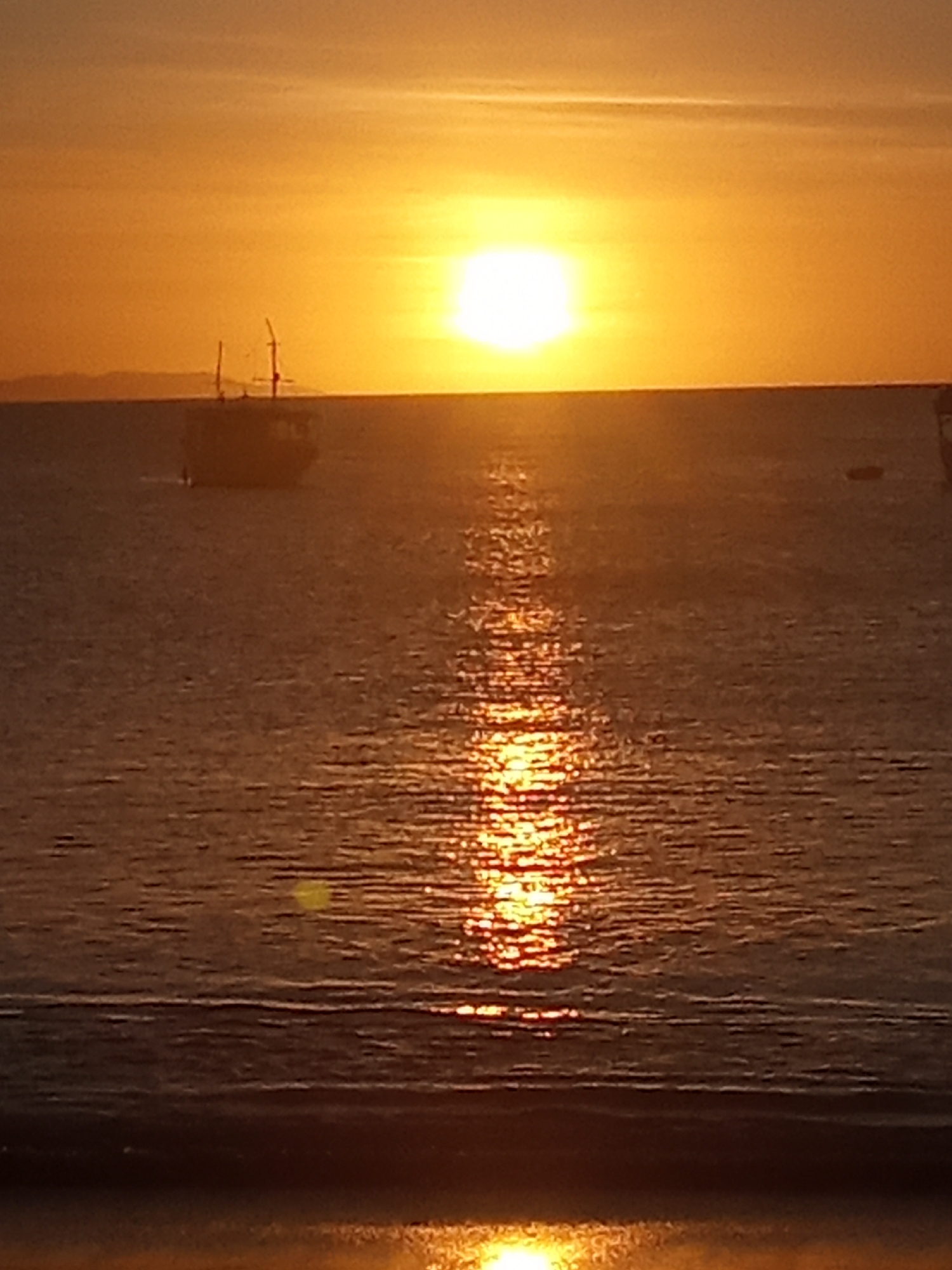 The image displays a maritime sunset or sunrise in Juan Griego, Venezuela. The bright, white-yellow sun is positioned slightly left of the frame's center on the horizon. The sky exhibits orange and yellow gradients, with faint cloud formations. A prominent, shimmering vertical reflection of golden light extends from the sun across the water to the foreground. A dark, two-masted vessel is silhouetted against the horizon, located to the left of the sun's reflection. A smaller, indistinct dark object, likely another boat, is visible further right and more distant on the water. Faint, dark landmasses are discernible on the distant horizon to the left. The immediate foreground features a dark, indistinct horizontal band. No people are present.