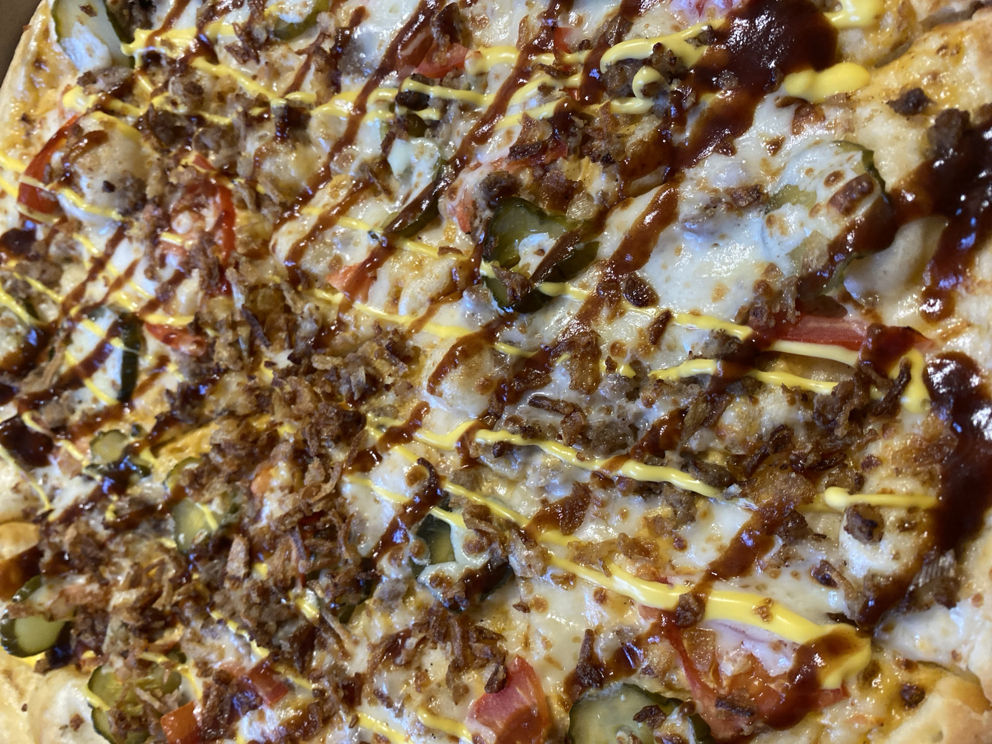 The image depicts a close-up view of a pizza, its toppings including cheese, ground meat, sliced tomatoes, pickles, fried onions, yellow mustard, and a dark brown sauce. It is not possible to determine the location of the image as Прикамский, Russia or any information about people, subjects, objects, actions, interactions, and the environment.