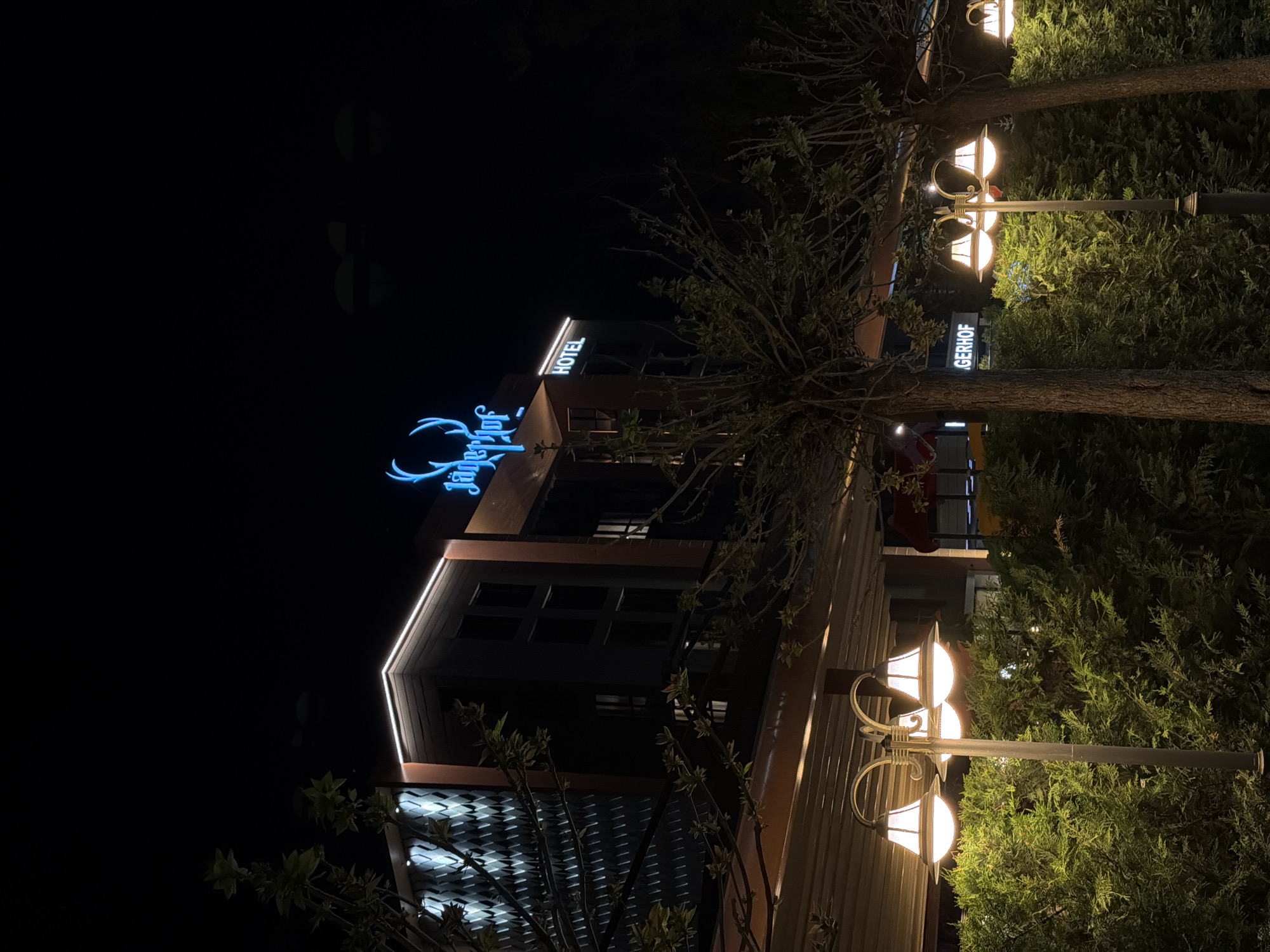 The image depicts a nighttime scene of a building with signage indicating it is the Jagerhof Hotel. The hotel signage features illuminated lettering and a deer antler emblem in a neon blue color. Above this, a smaller sign reads "HOTEL" in white letters. To the right of the hotel is the name "GERHOF", also illuminated. The building's design is modern, featuring a blend of materials like wood and dark metal. Streetlights with three upward-facing lamps are positioned in front of the building amidst lush greenery. The photo is taken at night, with a dark sky in the background. The location is Plovdiv, Bulgaria.