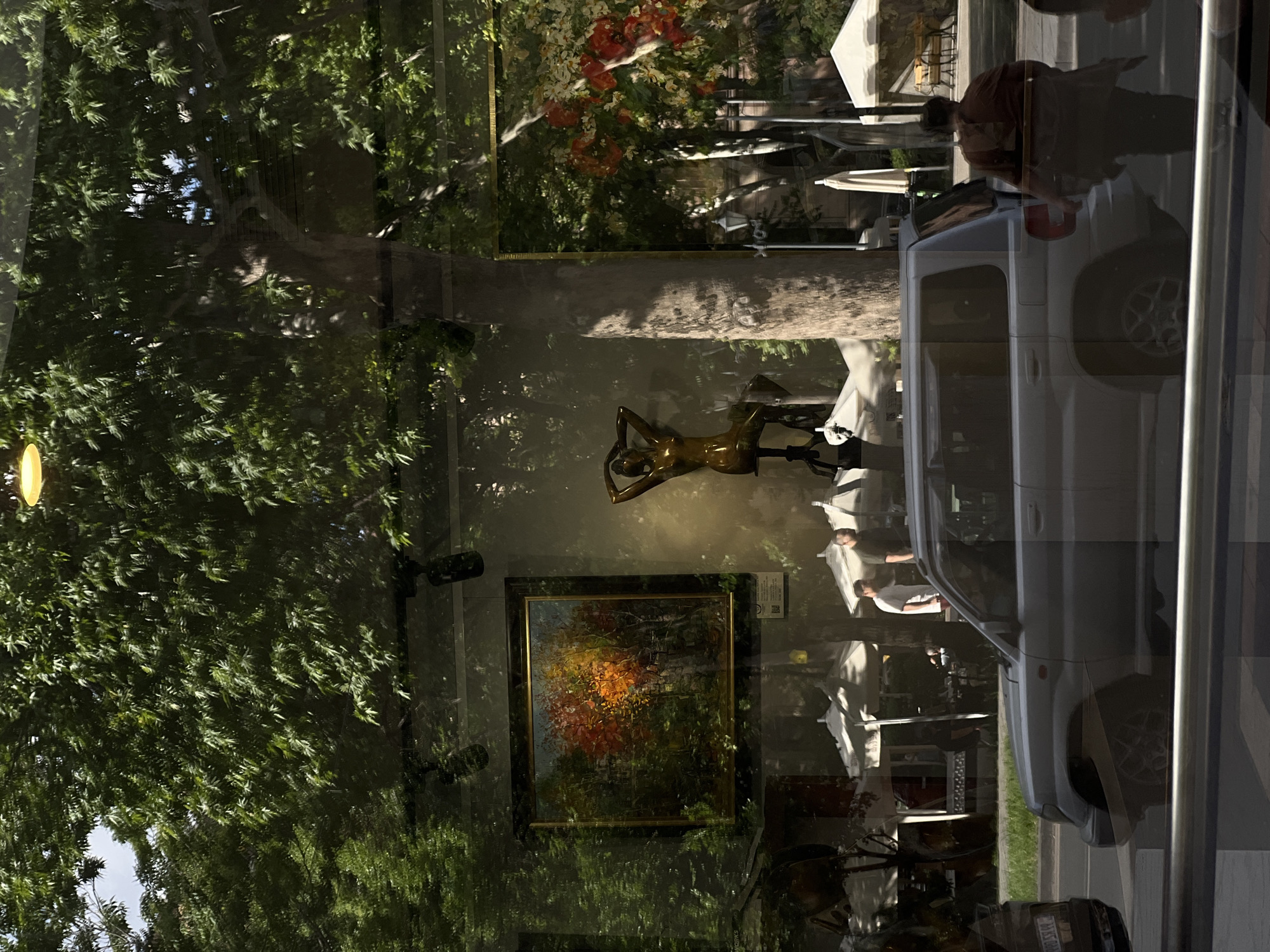 The image captures a layered scene through a large glass window, combining elements seen through the glass with strong reflections from outside. The photograph itself is oriented on its side, with dense green foliage from trees dominating the left and upper-left portions.
Through the glass, several items are visible inside what appears to be a shop or gallery. A golden or bronze-colored statue of a reclining female figure is centrally placed. Below it, a framed painting features an abstract landscape with vibrant autumn colors, predominantly reds, oranges, and deep greens. A warm, glowing light fixture is visible on the far left, and two dark surveillance cameras are mounted on an inner wall towards the right.
Strong reflections of an outdoor street scene are superimposed on the glass, particularly on the right and lower-right parts of the image. These reflections show a light-colored vehicle, possibly an SUV or van, parked along a street. A person wearing a dark shirt is partially visible near the vehicle, while another person in a white shirt stands further right, beneath one of several white outdoor umbrellas. These umbrellas, along with reflected tables and chairs, suggest the presence of an outdoor cafe or restaurant patio across the street. The street pavement and a curb are also visible in reflection. In the bottom right corner, a partial license plate of another vehicle is discernible, displaying "44 SS 99".
The lighting suggests a sunny daytime, with clear shadows visible in the reflections, indicating bright weather conditions. The setting is identified as Yerevan, Armenia. There are no discernible emotions conveyed by the individuals, and activity is limited to standing or waiting.