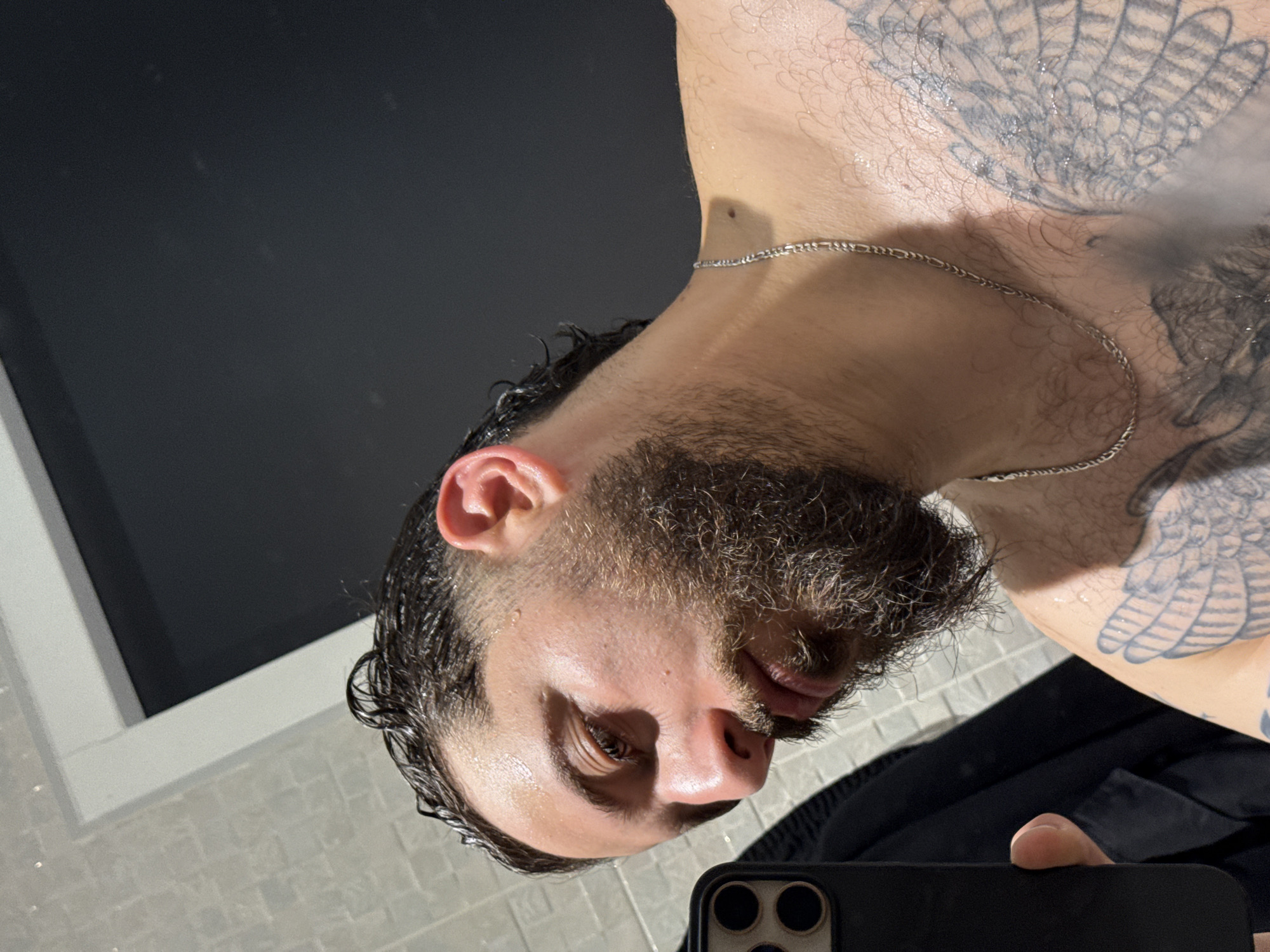 A male subject is depicted in a mirror selfie, seen from the mid-chest up. He has dark, wet, wavy hair and a full beard. Water droplets are visible on his skin, suggesting recent exposure to water. He wears a silver Figaro chain around his neck. Visible tattoos include large, feathered wing designs on his chest and right shoulder. His left hand is partially visible in the lower left corner, holding a black smartphone with a triple camera lens array, used to capture the image. The reflected environment behind him includes a light-colored, possibly white or light gray, tiled wall with grout lines on the left, and a dark, featureless surface framed by a white trim on the right, possibly a window or dark wall. A dark garment is also reflected behind the phone. The specific location within Strasbourg, France, is not discernable from the visual information provided in the image.
