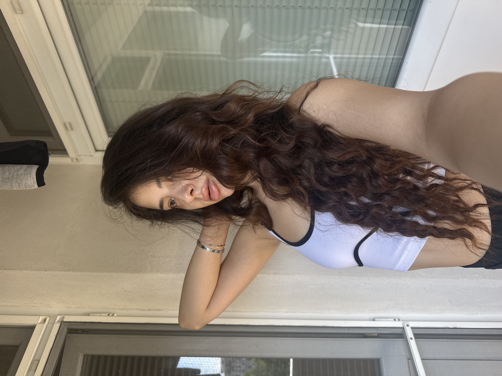 A young woman with long, dark, curly hair stands on a balcony, facing away from the camera and looking out a window. She is wearing a white crop top with black trim. Her right arm is raised and bent behind her head, with her hand resting on her neck. She has a silver bracelet on her left wrist. The background shows a textured wall, a window with blinds, and a screen door. The scene appears to be in a residential area, likely an apartment building, in Plovdiv, Bulgaria.