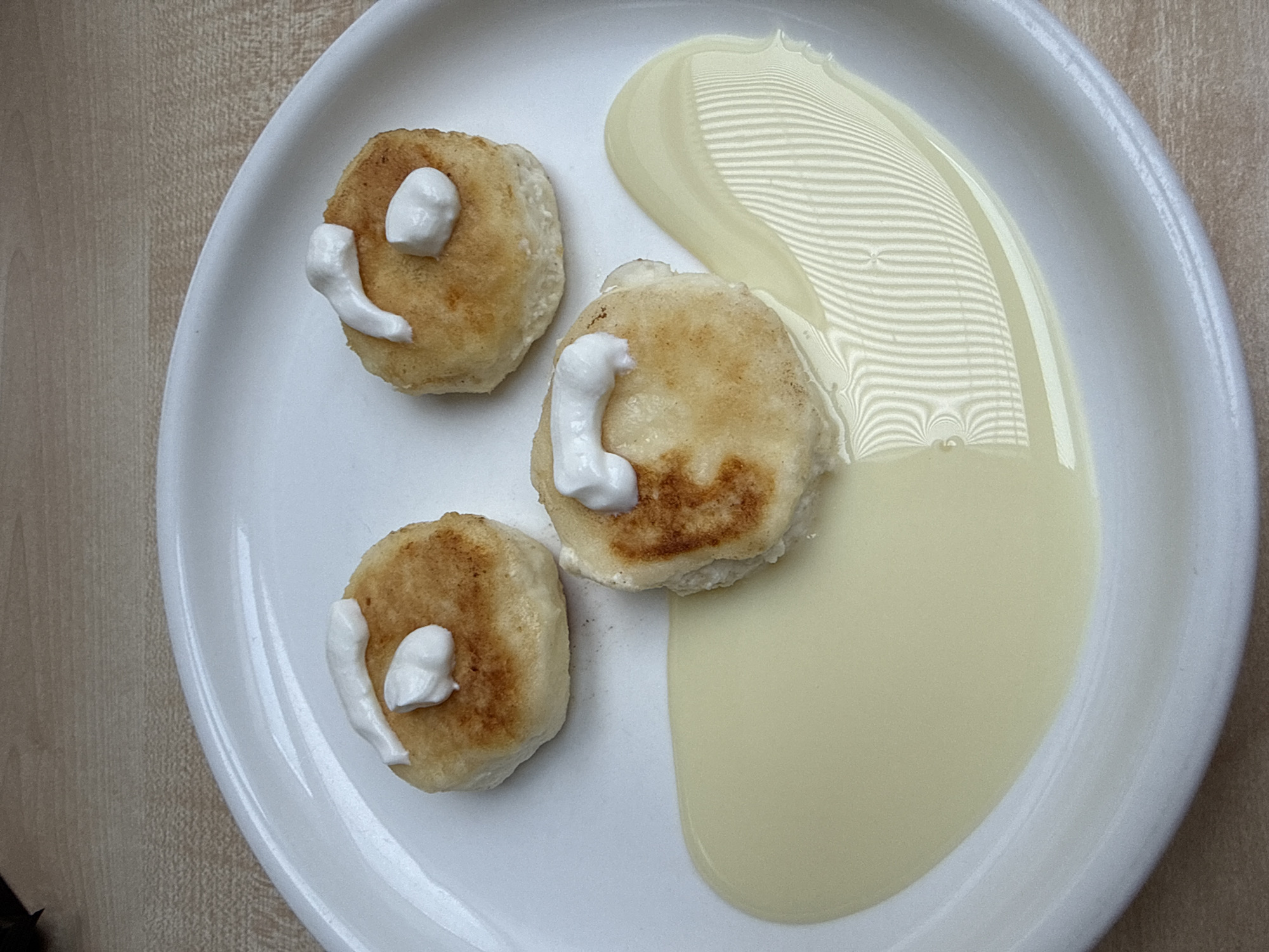 On a white plate are three small, round, golden-brown syrniki topped with dollops of white sour cream. Adjacent to the syrniki is a pool of light yellow, possibly condensed milk or custard sauce. The plate sits on a light brown wooden surface. The reflection of a patterned object, possibly window blinds, is visible on the surface of the sauce. The location is not discernible from the image.