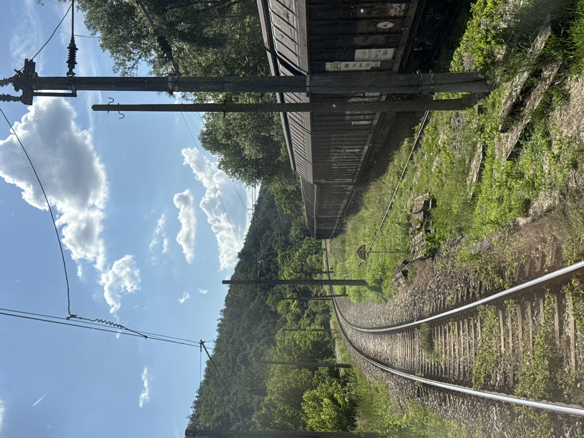 An outdoor, eye-level, medium shot captures a train track curving to the left, with a freight train running parallel on the right. The train, composed of multiple dark-colored cars with corrugated roofs, appears to be stationary. The tracks are surrounded by lush green grass and vegetation, with some scattered wooden ties in the foreground. Overhead, power lines stretch across the frame, supported by utility poles. A verdant, tree-covered hill rises in the background, under a bright blue sky dotted with fluffy white clouds. The scene is illuminated by natural sunlight, casting subtle shadows. No people are visible in the image.