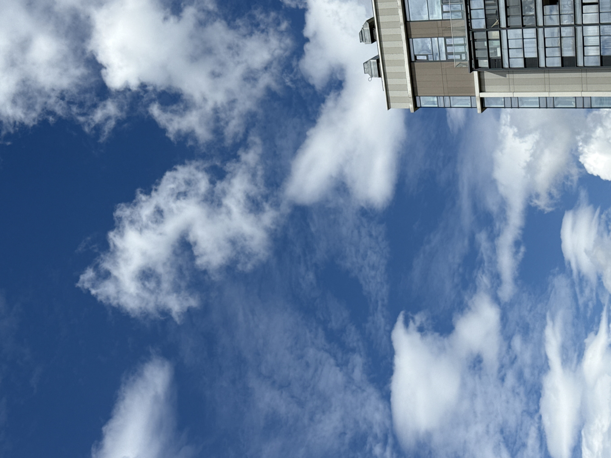 The image depicts the upper floors of a modern apartment building against a bright blue sky with scattered white clouds. The building features a mix of gray and beige facades, with large glass windows and balconies visible. Two ventilation units are situated on the rooftop. The angle of the shot suggests it was taken from below, looking upwards. There are no people or vehicles visible in the frame. Based on architectural style and common cloud formations, this scene is likely located in the city of Moscow, Russia.
