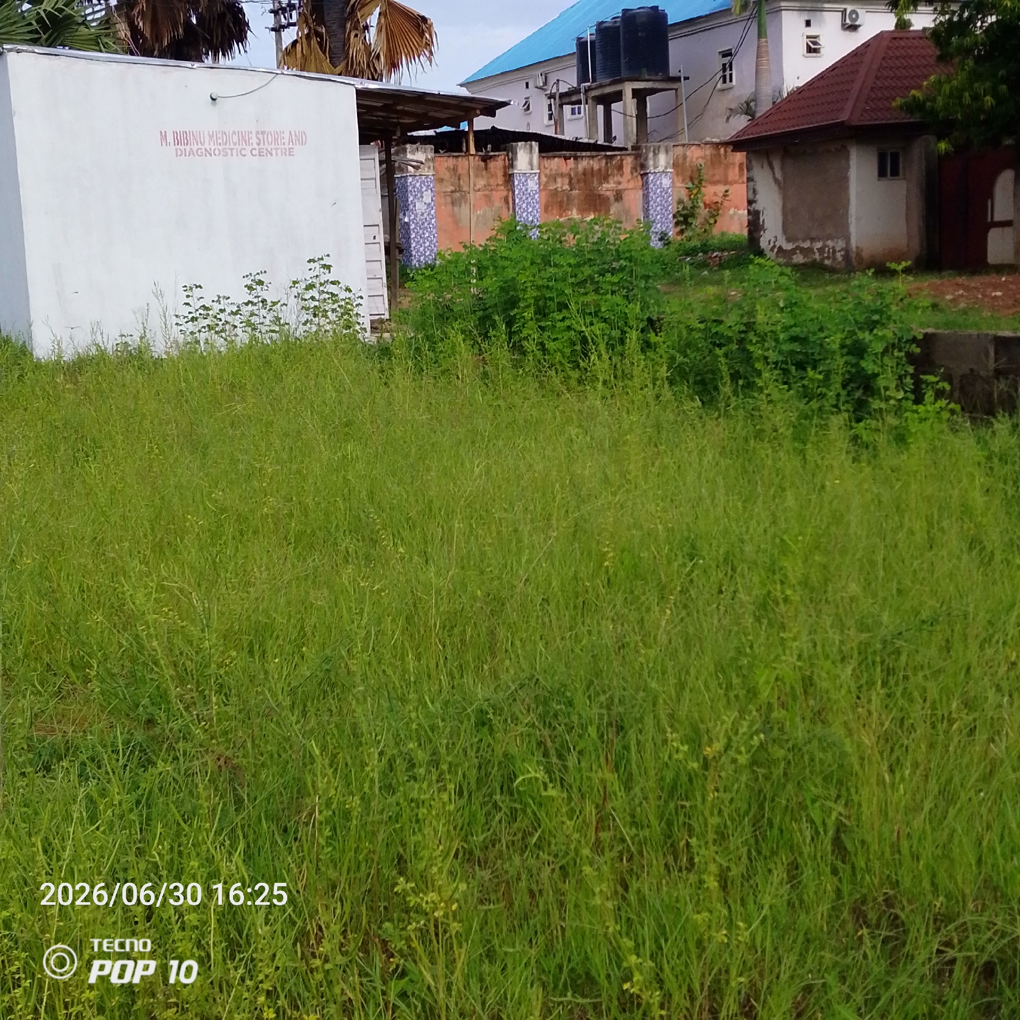 The image depicts an outdoor scene with a large, overgrown field of green grass in the foreground. In the background, several buildings and structures are visible. To the left, a white building with signage reads "M. BIBINU MEDICINE STORE AND DIAGNOSTIC CENTRE." Further back, there are other buildings, including one with a red tiled roof and another with a blue roof. A brick wall with decorative tile accents separates the immediate area from the background buildings. Several tall water tanks are also visible on top of one of the structures. The grass appears unkempt and is mixed with other plants. The overall impression is of a somewhat neglected area.
The image includes a timestamp in the bottom left corner: "2026/06/30 16:25". Below the timestamp, there is text and a logo that appears to be from a mobile phone manufacturer, "TECNO POP 10". The lighting suggests it is daytime, possibly late afternoon. The sky is overcast. There are no people visible in the image. The scene appears to be a residential or commercial area with undeveloped land.