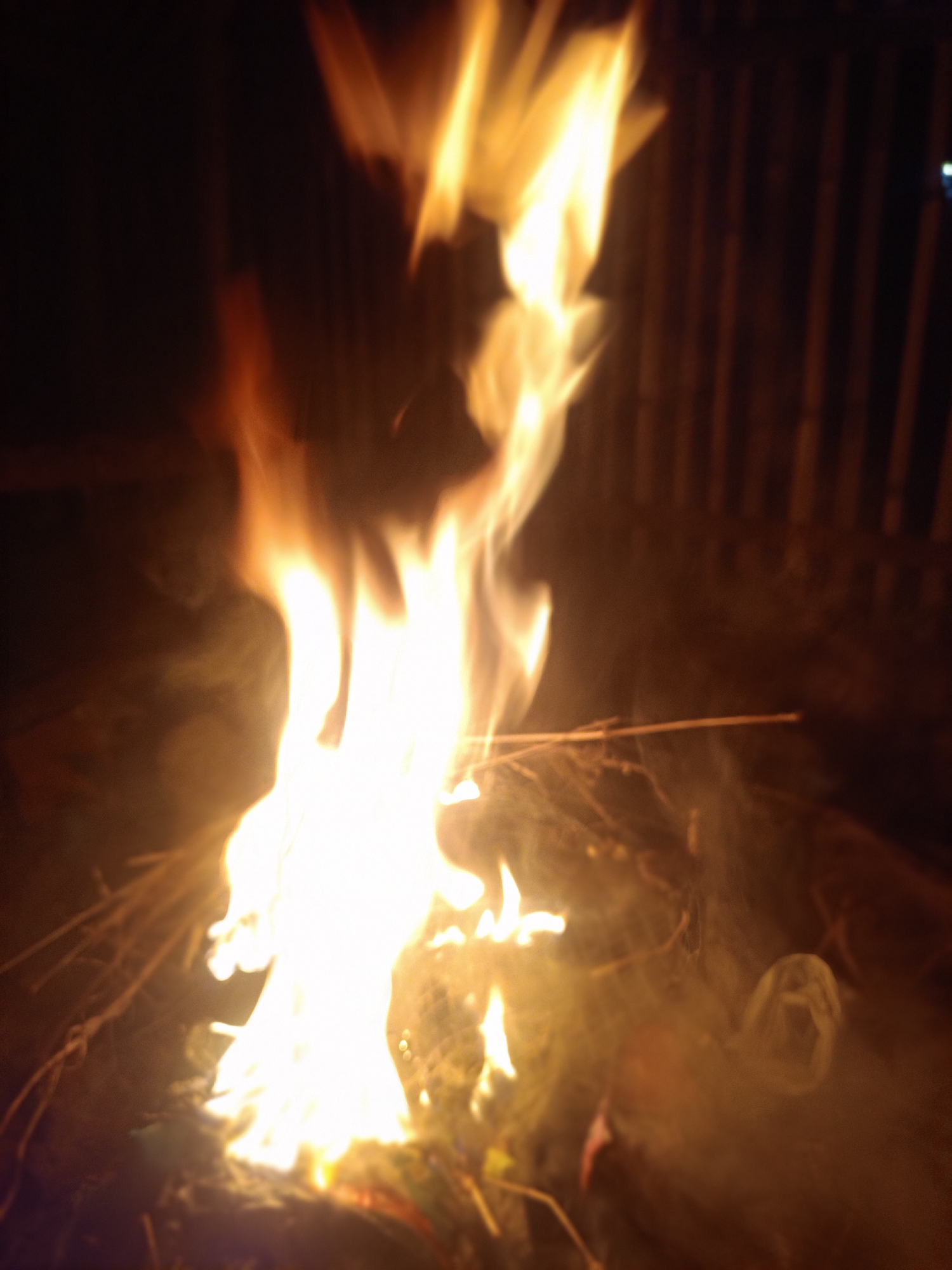 The image depicts a fire burning in the dark. The flames are tall and bright, illuminating the surrounding area. There are branches and debris beneath the flames, and smoke is visible rising into the air. In the blurred background, there is what appears to be a wall with vertical slats. The location is indicated as Buketteukuh, Indonesia, but there are no further details about people, interactions, or specific location within the city.