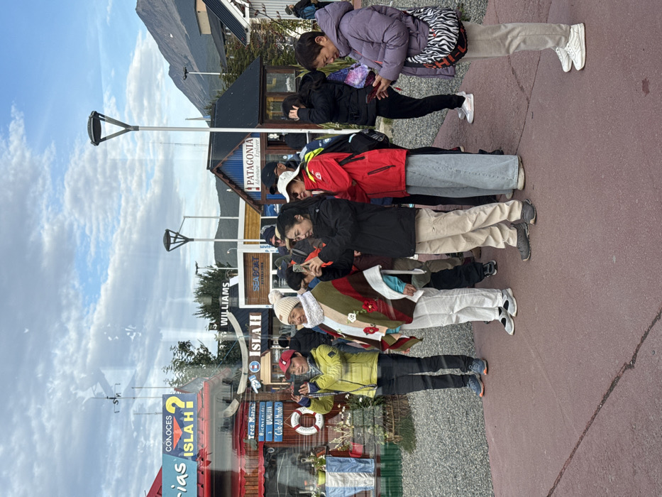 An outdoor scene in Ushuaia, Argentina, depicts a group of approximately ten individuals, predominantly East Asian in appearance, standing on a reddish-brown paved surface. The setting appears to be a waterfront tourist area, characterized by several small, colorful buildings in the mid-ground, including one red structure displaying "CONOCES ISLA H?", another featuring "WILLIAMS", a blue building with "SEABOAT", and a blue and white striped building labeled "PATAGONIA Adventure Explorer." Distant, snow-capped mountains rise under a partially cloudy sky with visible patches of blue.
In the immediate foreground, a reflection on a glass surface partially obscures the view. Among the people, two individuals (a person in a neon yellow/green jacket and a person in a black jacket) are actively holding black smartphones horizontally, seemingly capturing photos or videos of the surroundings. Another individual, an older woman in a lavender puffer jacket carrying a zebra-striped bag, is focused on a white smartphone held vertically. Other members of the group are dressed in various casual outdoor attire, including puffer jackets, ponchos, and hats, observing the scene. One person wears a red jacket and light blue wide-leg jeans. The ground beyond the paved area is covered with light-colored gravel. Two modern, dark-colored streetlights with distinctive triangular heads are visible in the background.