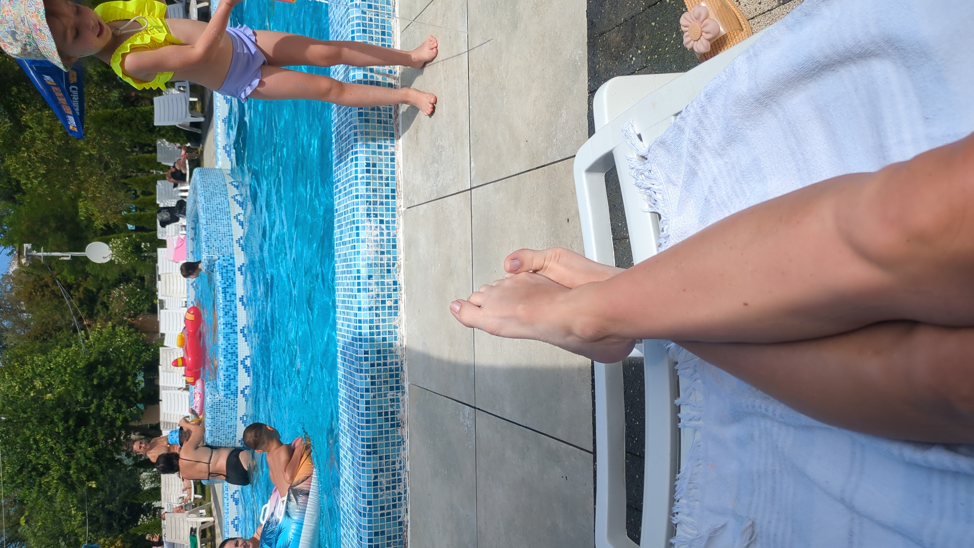 The image depicts a poolside scene in Vadul lui Voda, Moldova. In the foreground, a person's bare legs and feet are crossed, resting on a white lounge chair covered with a white towel. The feet appear to have light-colored nail polish. Behind the lounge chair, the edge of a swimming pool is visible, lined with blue and white mosaic tiles. The pool water is a vibrant blue, and several people are enjoying it. In the mid-ground, a woman in a black bikini is in the pool, facing away from the camera, with her arm around a child wearing a blue hat. A red inflatable toy in the shape of an airplane is also in the pool. To the right of the pool, a young girl in a yellow top and purple bottoms stands near the edge, wearing a patterned sun hat. Further in the background, trees and lounge chairs are visible, suggesting an outdoor recreational area. The overall impression is a sunny day at a pool, likely a resort or public swimming facility.
