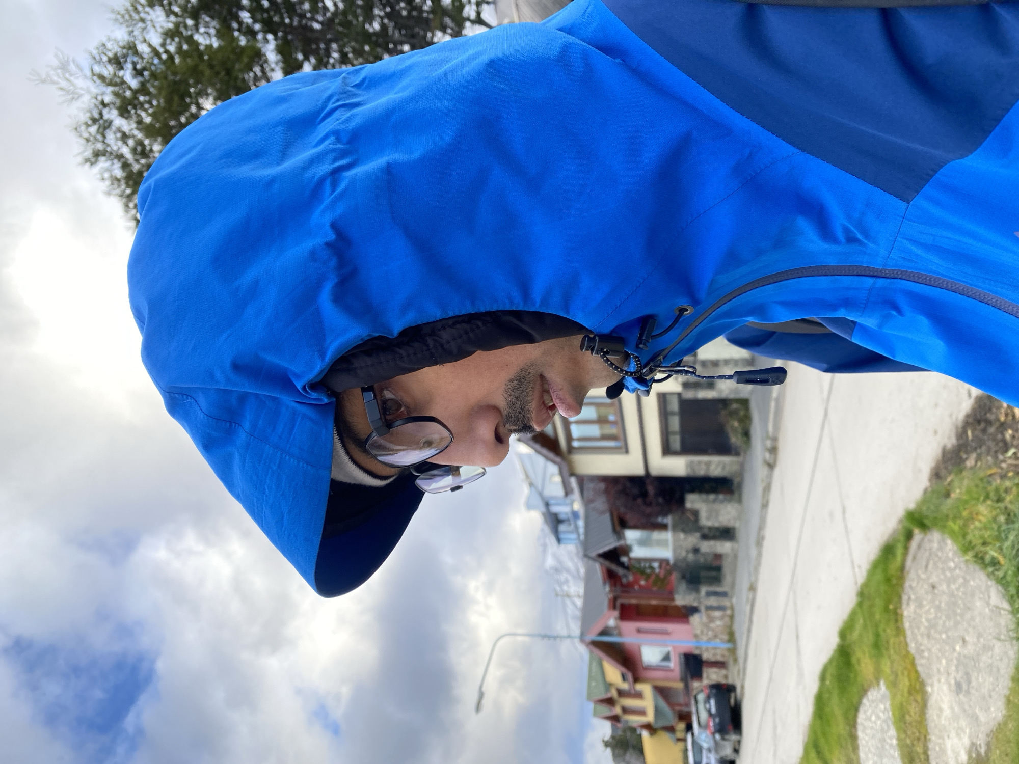 A man wearing glasses and a blue hooded jacket is standing on a sidewalk, with his head turned towards the camera. The background includes houses, a street, and a cloudy sky. The location is Ushuaia, Argentina.