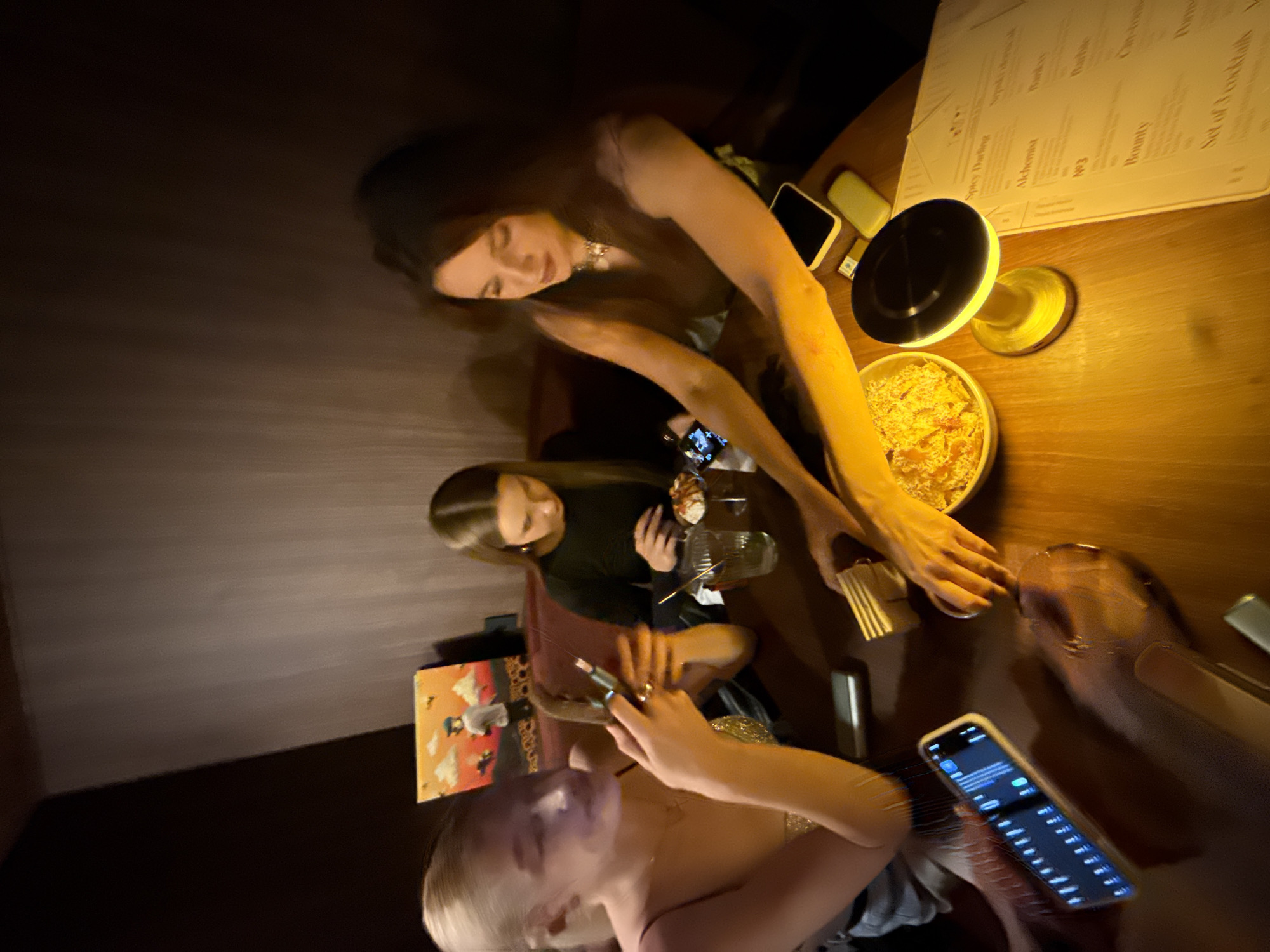 Three women are seated at a table in a dimly lit restaurant. The table is covered with food and drinks, including a bowl of chips, a portable light fixture, and a menu. One woman is looking at her phone. Belgrade, Serbia.