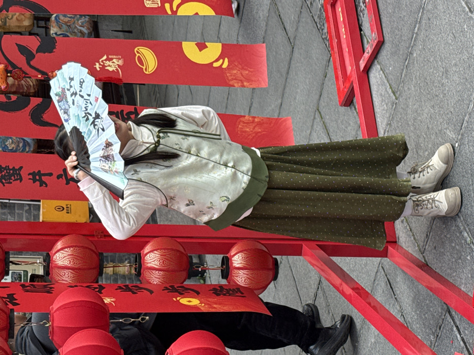 The image shows a person standing amidst vibrant red decorations, likely at an outdoor event or celebration. The photograph is oriented sideways, with the person standing vertically on the right side of the frame as it's presented.
The main subject is a person, likely a young woman, seen from the waist up, facing slightly away from the camera. She is holding a light blue, intricately decorated folding fan in her right hand, partially obscuring her face. Her left arm is by her side. She is wearing a light-colored, possibly off-white or very light green, long-sleeved top, over which she has a traditional-style vest. The vest is a pale greenish-cream color with subtle patterns, possibly embroidered butterflies or flowers, and features traditional Chinese frog closures at the neck. A darker green trim is visible at the bottom of the vest. She wears a long, flowing skirt in a dark olive green color with small, light-colored polka dots. On her feet, she wears light-colored, possibly off-white or beige, lace-up boots with thick soles.
The background and foreground are dominated by red structures and decorations. To her left (viewer's right in the rotated image) is a red wooden frame or stand. Hanging from various points on these structures are numerous red banners or scrolls. These banners feature black Chinese calligraphy and golden symbols, including what appears to be a gold ingot (元宝 - yuánbǎo), a traditional symbol of prosperity, and a traditional Chinese coin symbol. Some banners have a textured, slightly worn appearance. Below the person's eye level, several red lanterns are visible, some with golden intricate patterns. In the bottom left corner, part of another person's legs and black shoes are visible, suggesting more people are present.
The ground is paved with grey stones, indicating an outdoor setting, possibly a street or courtyard. The overall atmosphere appears festive and cultural, likely related to a Chinese celebration such as Chinese New Year due to the prevalent red color, lanterns, and traditional symbols.
Visible text includes Chinese characters on the red banners. Some characters are clearly visible in black, such as "新年好" (Xīnnián hǎo - Happy New Year) and others that are part of couplets or blessings. The small golden word "招財" (zhāocái - attracting wealth) is also visible on one of the banners, above a gold ingot symbol.