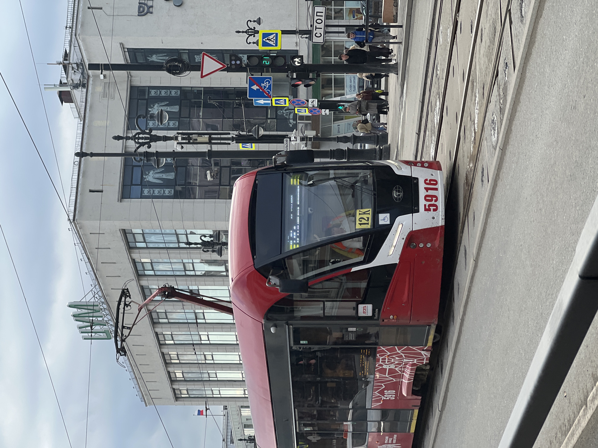 A red and white tram, number 5916 and route 12K, is depicted in the city of Perm, Russia. It is on tram tracks near a pedestrian crosswalk. The background includes buildings, streetlights, and traffic signs, including a "Stop" sign and pedestrian crossing signs. People are seen crossing the street in the background.