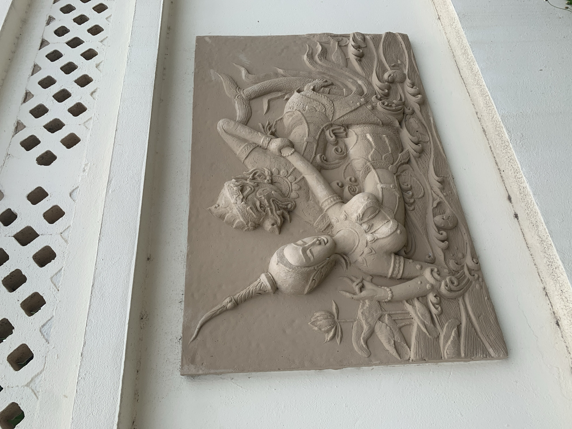 A bas-relief sculpture depicting a figure of divinity adorns a white wall in Baan Bo Phut, Thailand. The rectangular sculpture is executed in a light brown, unpainted material, possibly stone or cement. It showcases an intricate design with a central figure in a dynamic, bent posture, possibly indicating movement or dance. Elaborate details depict the figure's ornamentation, headdress, and flowing elements. The surrounding composition features foliage and possibly other figures or mythological elements, all carved in the same low-relief style. The adjacent wall is painted white and features a decorative lattice pattern. The environment suggests an outdoor setting, likely part of a temple, shrine, or another religious or cultural site in Baan Bo Phut.
