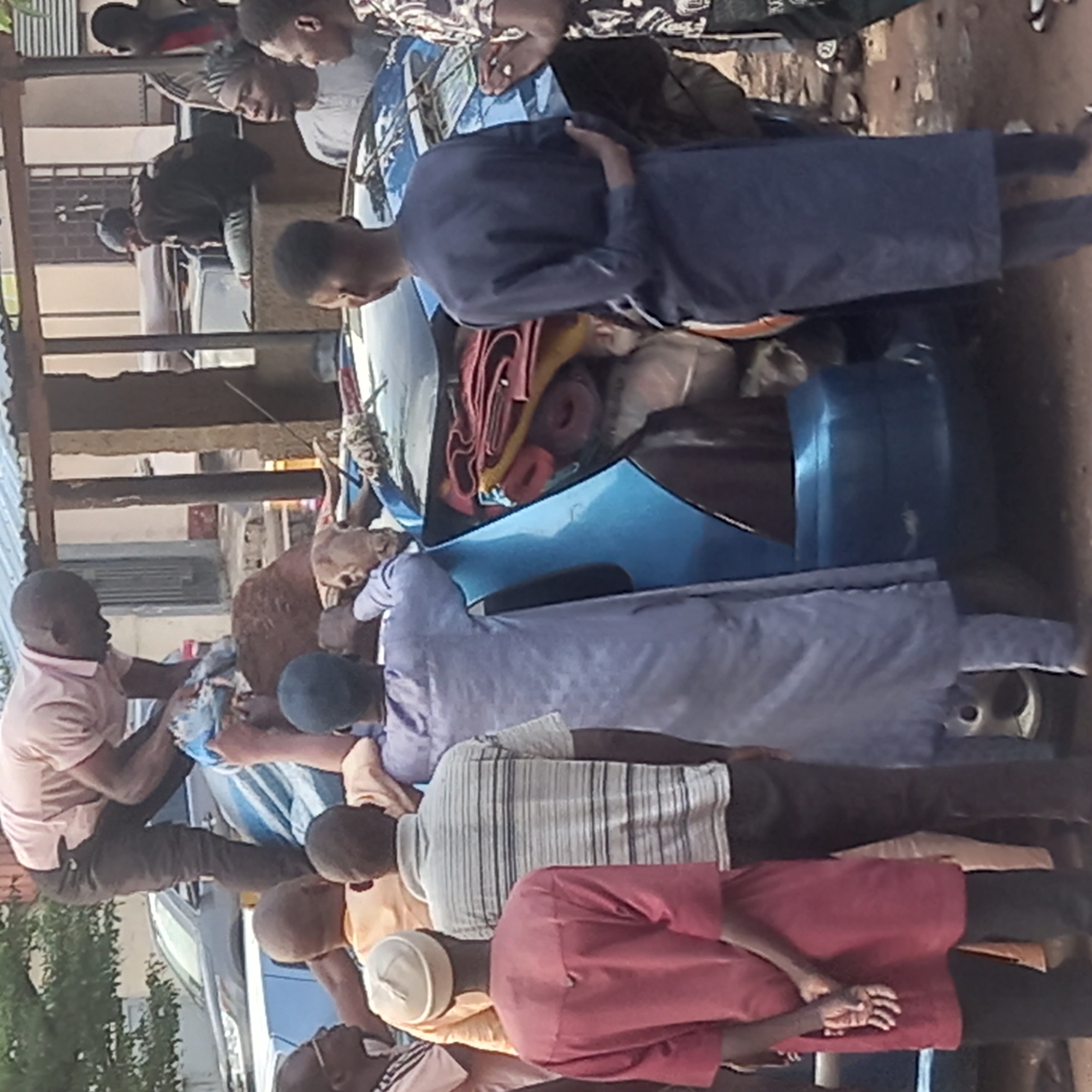 The image shows a group of men gathered around a blue car heavily loaded with various items, including what appears to be a goat carcass. The men are dressed in a variety of clothing styles, some in traditional attire. The scene appears to be outdoors, possibly in a residential or commercial area. The location is Dadi Riba, Nigeria.