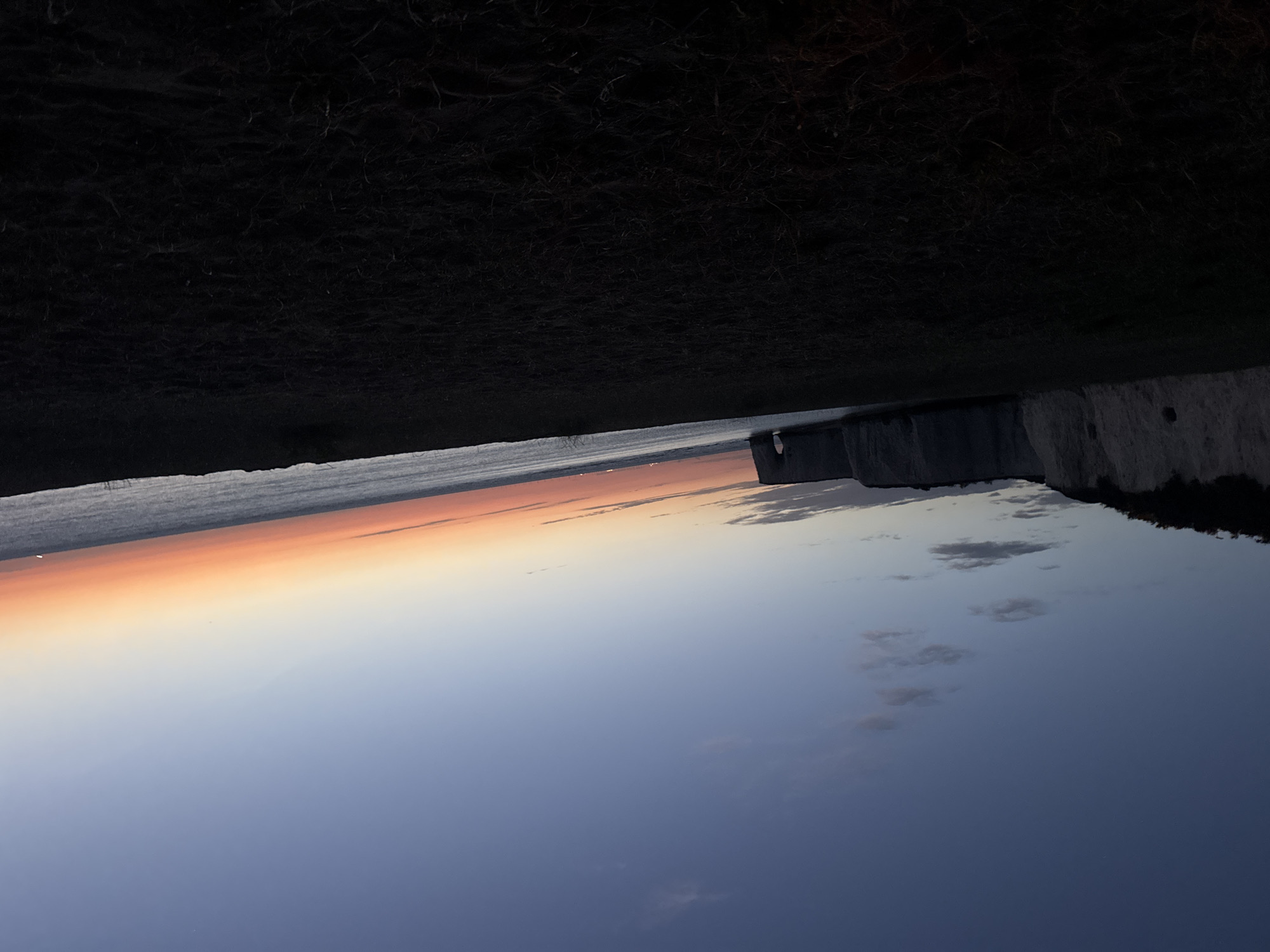 This media file captures a dramatic seascape at sunset. The scene is dominated by a dark, textured foreground that appears to be grassy terrain, possibly the top of a cliff. Below this, a vast expanse of sky transitions through a gradient of colors, from a fiery orange and pink at the horizon to a deep, serene blue. This suggests it is late evening, during or just after sunset.
The horizon is marked by the distinct white cliffs characteristic of the Thanet coastline in the United Kingdom. The cliffs are rugged and imposing, with some vegetation visible on their tops. Below the cliffs, the sea is calm and reflects the colors of the sky. A few scattered clouds are present in the sky, adding texture to the twilight hues.
There are no people or specific objects clearly discernible in the image. The overall mood is one of tranquility and natural beauty, highlighting the majestic interplay of light, land, and sea during a sunset. The orientation of the image is rotated, with the sky appearing at the bottom and the land at the top.