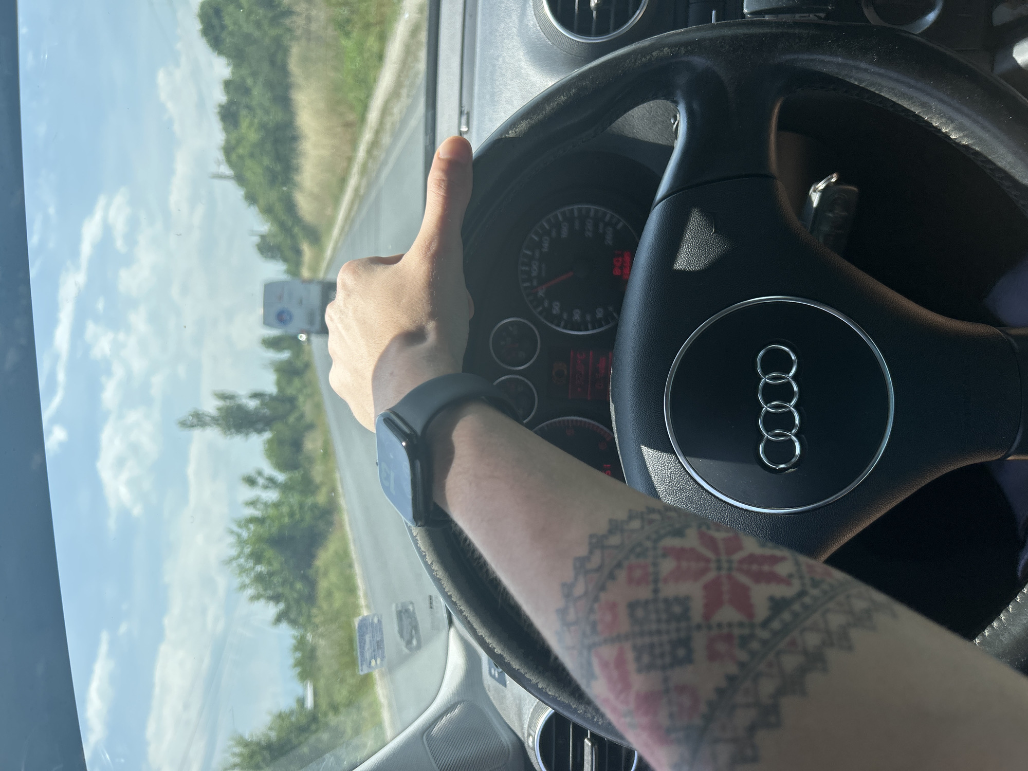 The image shows a close-up, first-person perspective from inside a car, featuring a person's right arm and hand giving a thumbs-up gesture. A dark smartwatch is visible on the wrist. The forearm is extensively tattooed with intricate red and black geometric patterns, characteristic of traditional Eastern European embroidery.
The setting is an Audi car interior, clearly indicated by the four-ring logo on the black leather steering wheel. Behind the hand, parts of the car's dashboard and instrument cluster are visible, including a speedometer showing a speed of approximately 80-90 km/h, a tachometer, and some illuminated warning lights, one of which is red. Car air vents are also present in the dashboard.
Through the car's window, a bright, sunny outdoor scene is visible. The sky is largely clear with white clouds. Green trees and foliage line a road that stretches into the distance, suggesting a drive through a rural or highway landscape. The time of day is daytime, with strong sunlight. The thumbs-up gesture conveys a positive, approving, or content emotion, likely related to the journey. The provided location context is Dalgo Pole, Bulgaria.