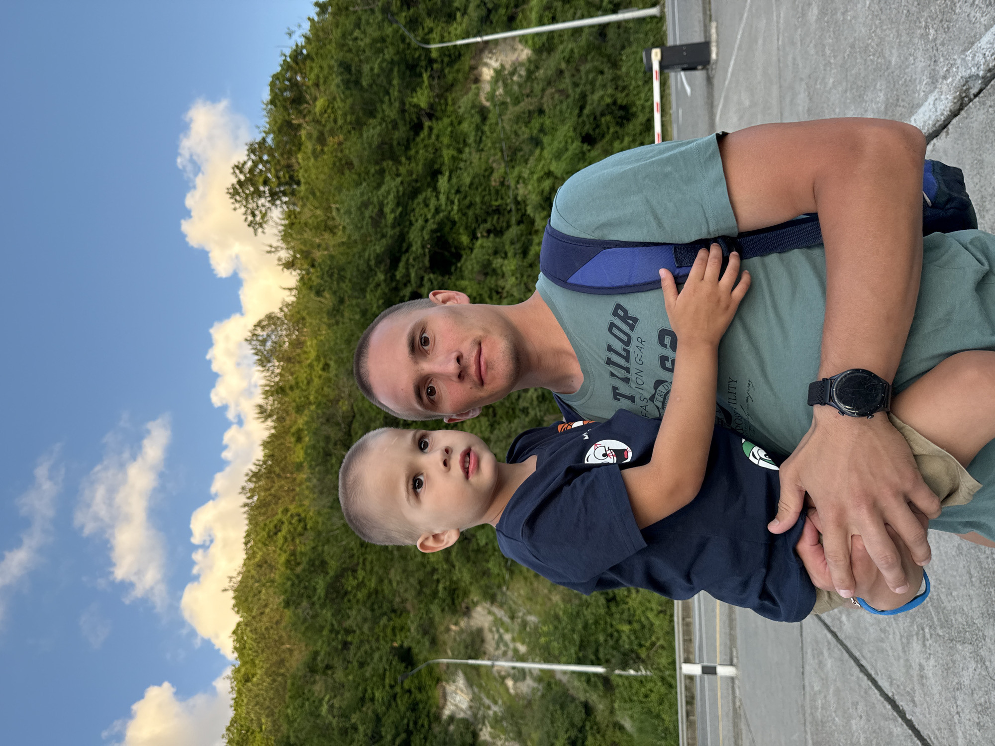 A man is holding a young boy in front of a lush green hillside under a blue sky with scattered white clouds. The man is wearing a teal t-shirt with a backpack over his shoulder, and a watch on his left wrist. The boy is wearing a dark blue t-shirt. They appear to be outdoors, possibly in a scenic area. In the background, there is a road, a barrier, and a lamppost. The location is specified as Nebug, Russia.