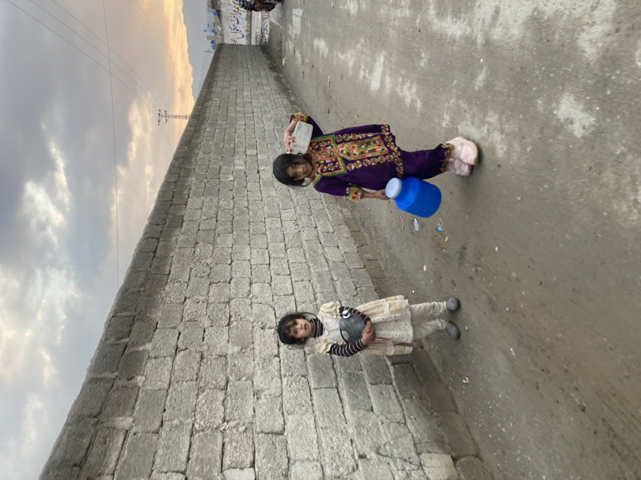 Two young girls are depicted standing on an unpaved dirt path in Quetta City Tehsil, Pakistan, adjacent to a long, grey concrete block wall. The girl on the left, wearing a light-colored dress over a striped shirt and light pants, holds a grey metallic pot or kettle in both hands. The girl on the right, dressed in a purple traditional attire with extensive embroidery, carries a blue plastic jerrycan with a white lid in her left hand and a clear plastic bottle on her right shoulder. Both girls face the viewer. The sky above is cloudy with faint hues of orange light, suggesting either sunrise or sunset. In the background, beyond the wall, a utility pole, distant mountains, and a partially visible motorcycle are present. A white sign with dark text, likely Urdu, is visible on the wall in the midground.