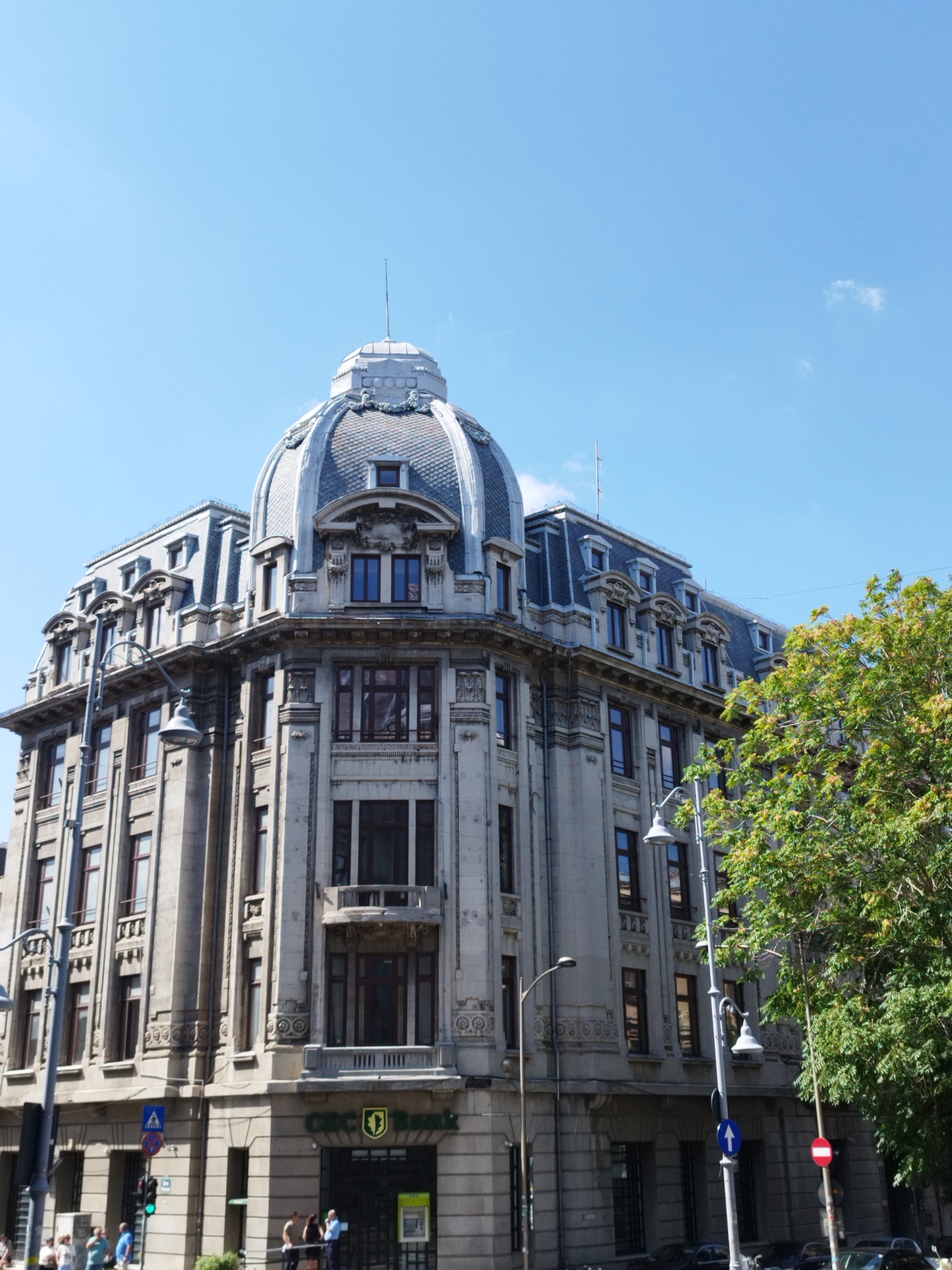 The image shows a grand, stone building with a large dome, a mansard roof, and ornate architectural details. The building is located in Bucharest, Romania, as evidenced by the "BRD - Groupe Société Générale" logo visible on a sign. The sky is clear blue, suggesting a sunny day. A large, leafy tree is on the right side of the frame, and a street lamp is visible in the foreground. Several people are seen on the sidewalk in front of the building, and cars are parked along the street. Traffic lights and street signs are also present, indicating an urban setting. The building appears to be a bank, likely a branch of BRD.