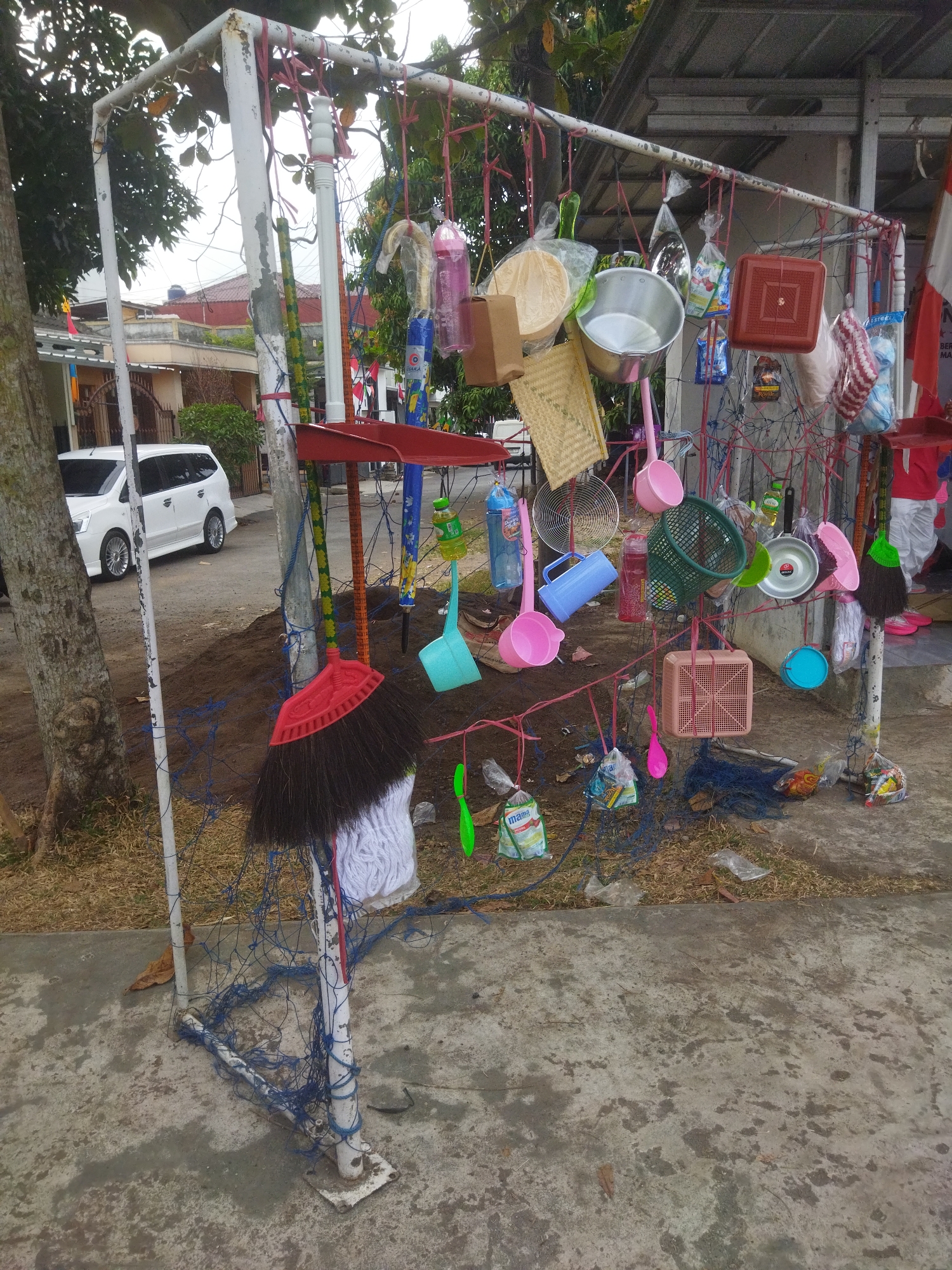 The image depicts a makeshift display of household cleaning supplies and kitchenware, likely for sale, outside a building. The items are hung from a white metal frame, possibly a repurposed goalpost, using various cords and strings. The display includes brooms, dustpans, buckets, basins, and other plastic containers. Some items are packaged in plastic bags. The scene is located outdoors on a paved surface next to a dirt area and some grass. A white car is parked on the road in the background, and a building with a corrugated metal roof is visible. The overall impression is of a humble, possibly informal, marketplace in a residential or commercial area. The specific location is identified as Banjar, Indonesia, based on external information.