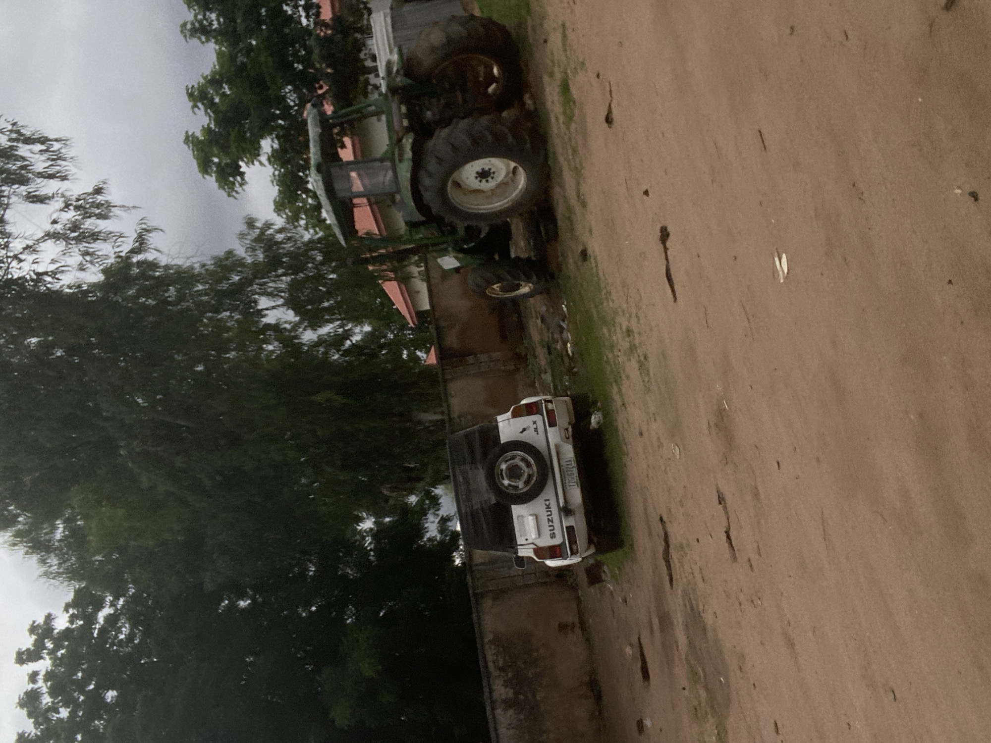 The image depicts an outdoor scene on a cloudy day, likely in Gbaza, Nigeria. In the foreground, a dirt ground area is visible with scattered debris, including what appear to be small pieces of wood or plant matter.
To the left, a white Suzuki SUV is parked facing away from the viewer. The vehicle has a spare tire mounted on its rear door and a license plate with the inscription "TZG 672 AA". The Suzuki is positioned in front of a rough, textured concrete wall that extends across the middle ground of the image.
To the right of the Suzuki and further back, a large green tractor is parked. The tractor appears to be in a field or a yard, with tall trees and foliage forming a dark green backdrop. The tractor has large, robust tires and a cab structure. Behind the tractor and the wall, buildings with red roofs and white walls are partially visible, suggesting a residential or industrial area. The overall environment is natural and somewhat unkempt, with the presence of both vehicles suggesting a functional, possibly rural or semi-rural, setting.