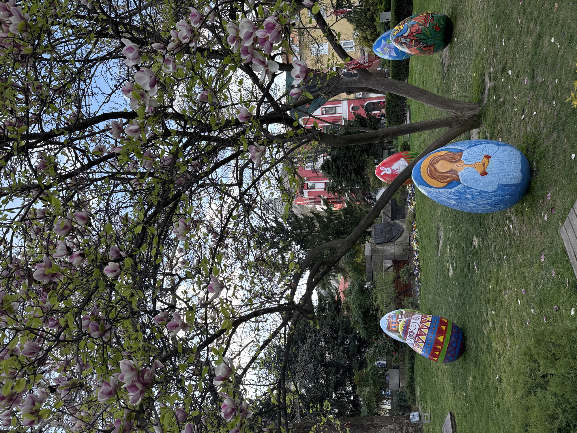 The image depicts a park setting in Plovdiv, Bulgaria. In the foreground, a magnolia tree with blossoms in shades of pink and white dominates the left side. Scattered around the base of the tree and on the grass are several large, decorated Easter eggs. Each egg is painted with different motifs: one features a woman with a trumpet, another a rabbit, and others with abstract and floral designs. In the background, buildings with red facades are visible. The setting suggests a festive display during the Easter season in an urban park environment.