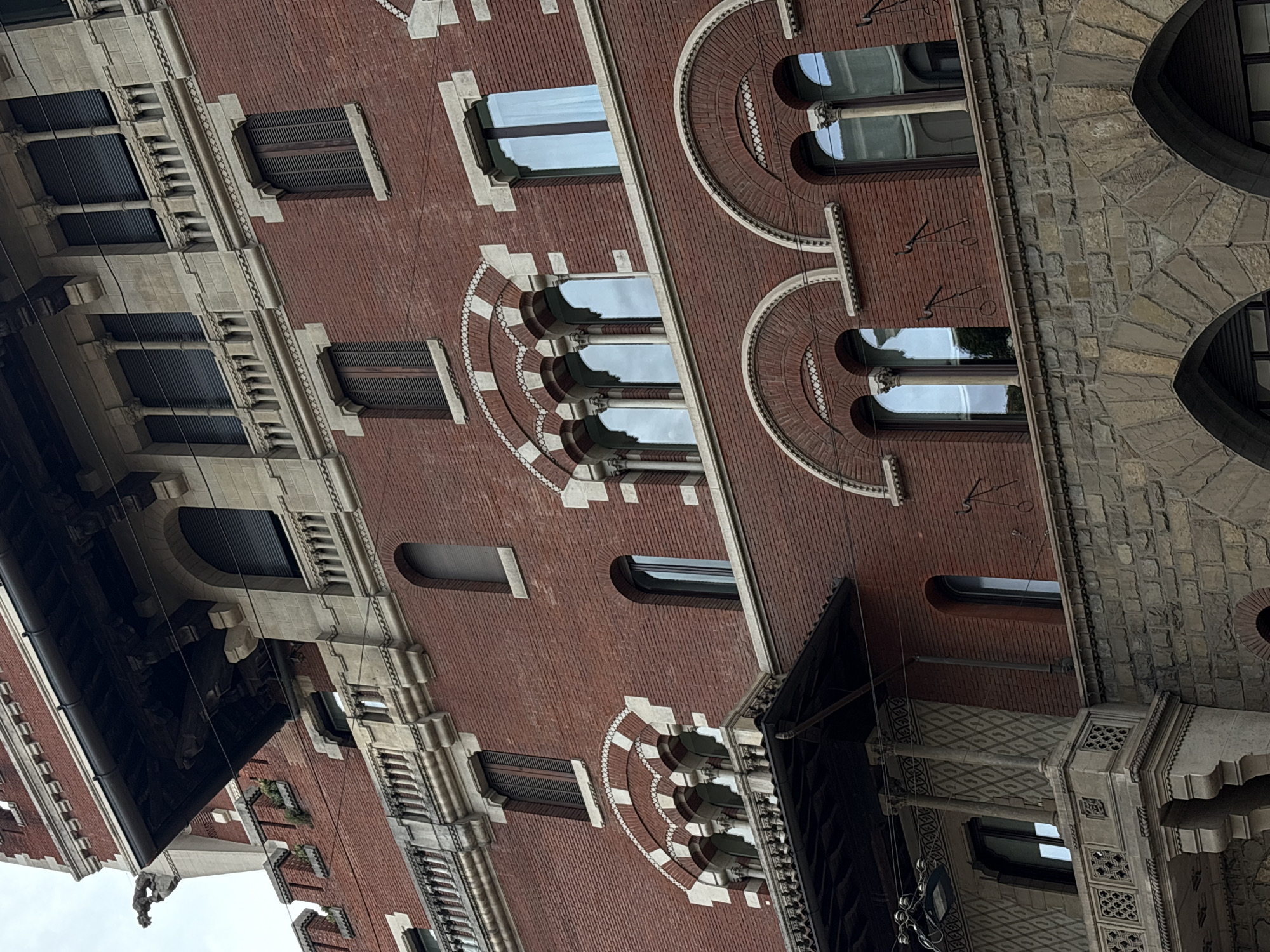 The image depicts a building facade in Milan, Italy, characterized by red brick and stone construction. Architectural details include arched windows, columns, and decorative masonry. The building's facade is multi-storied, with varying window styles and ornamental features along the upper levels. The environment is urban, with the building likely part of a larger streetscape.